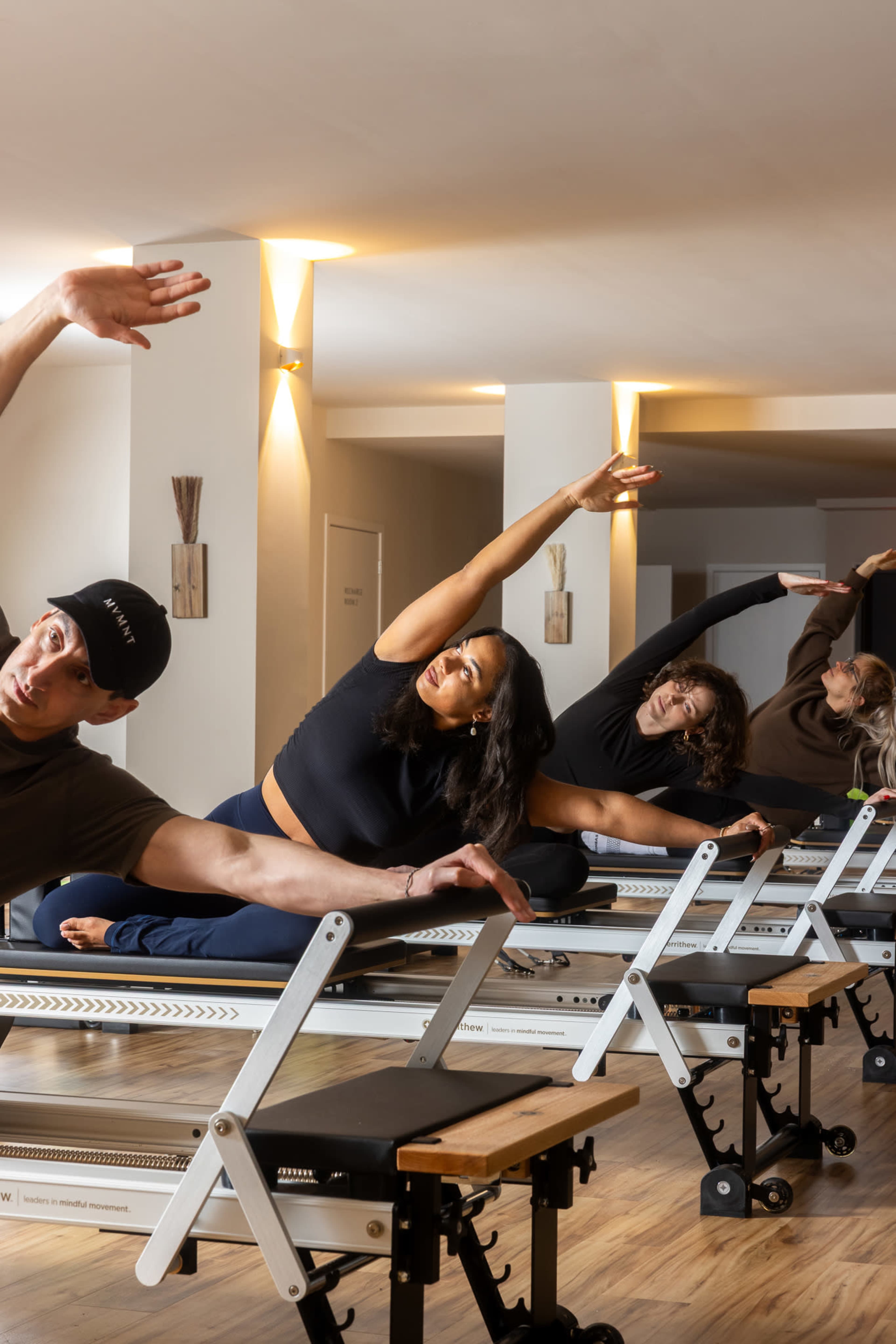 A group of four individuals is engaged in a Pilates class, performing side stretches on reformer machines in a brightly lit studio.