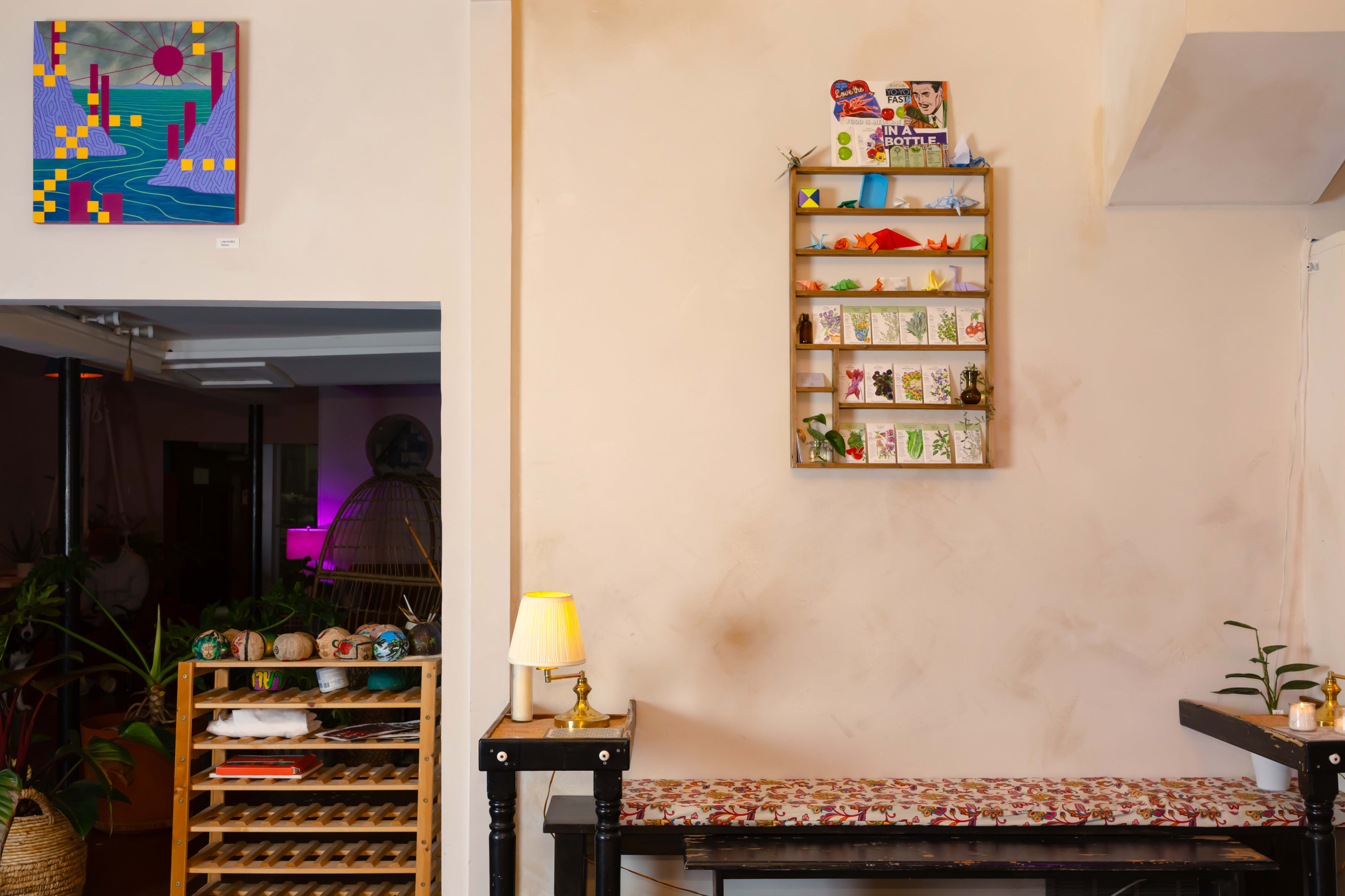 The image shows a cozy interior featuring a wall shelf with various small items, a bench with patterned fabric, and a lamp on a black table, alongside art on the wall.