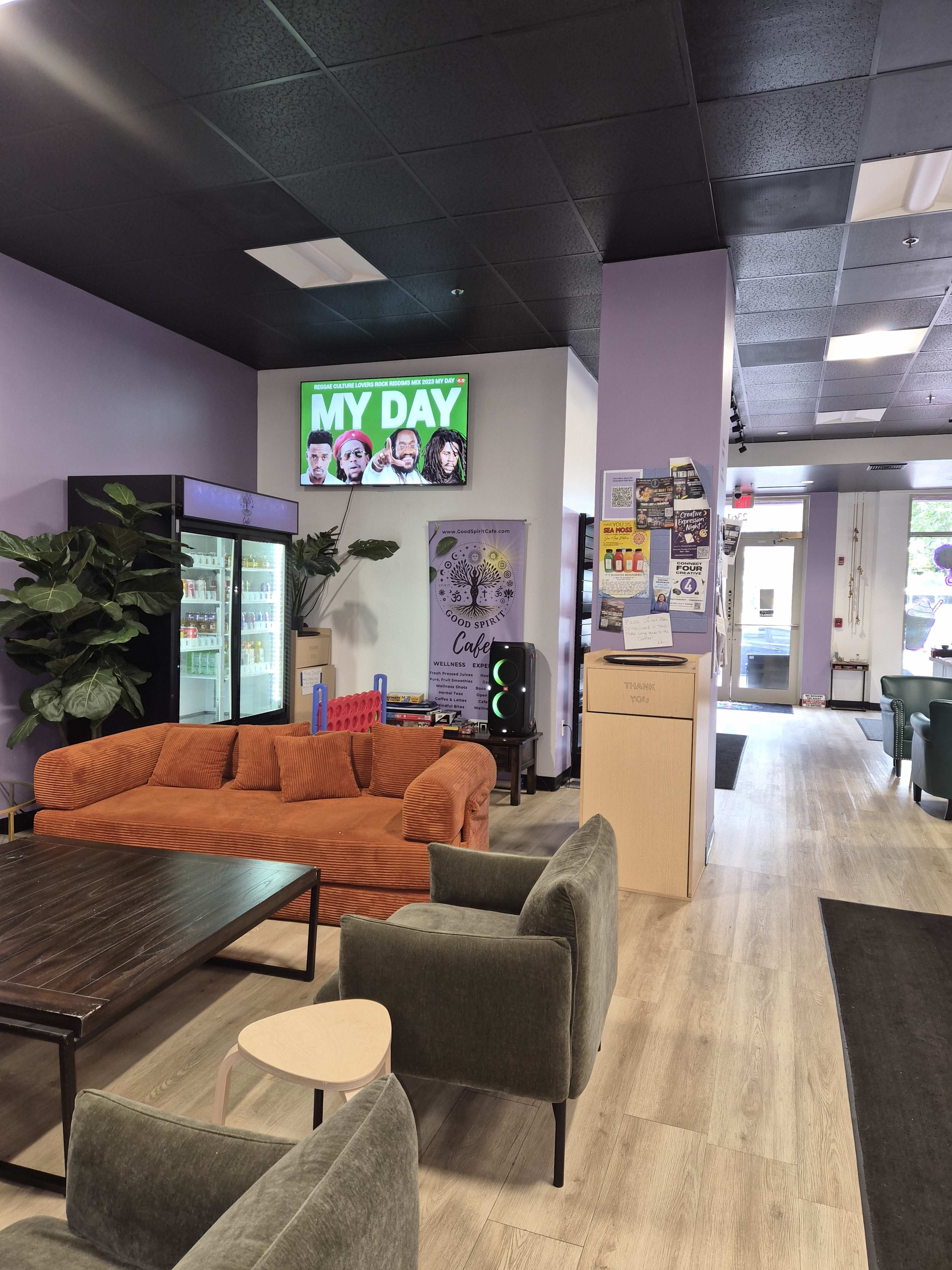 The image shows an interior space featuring a seating area with an orange couch, a wooden table, and various promotional materials on the walls, along with a digital screen displaying "MY DAY."