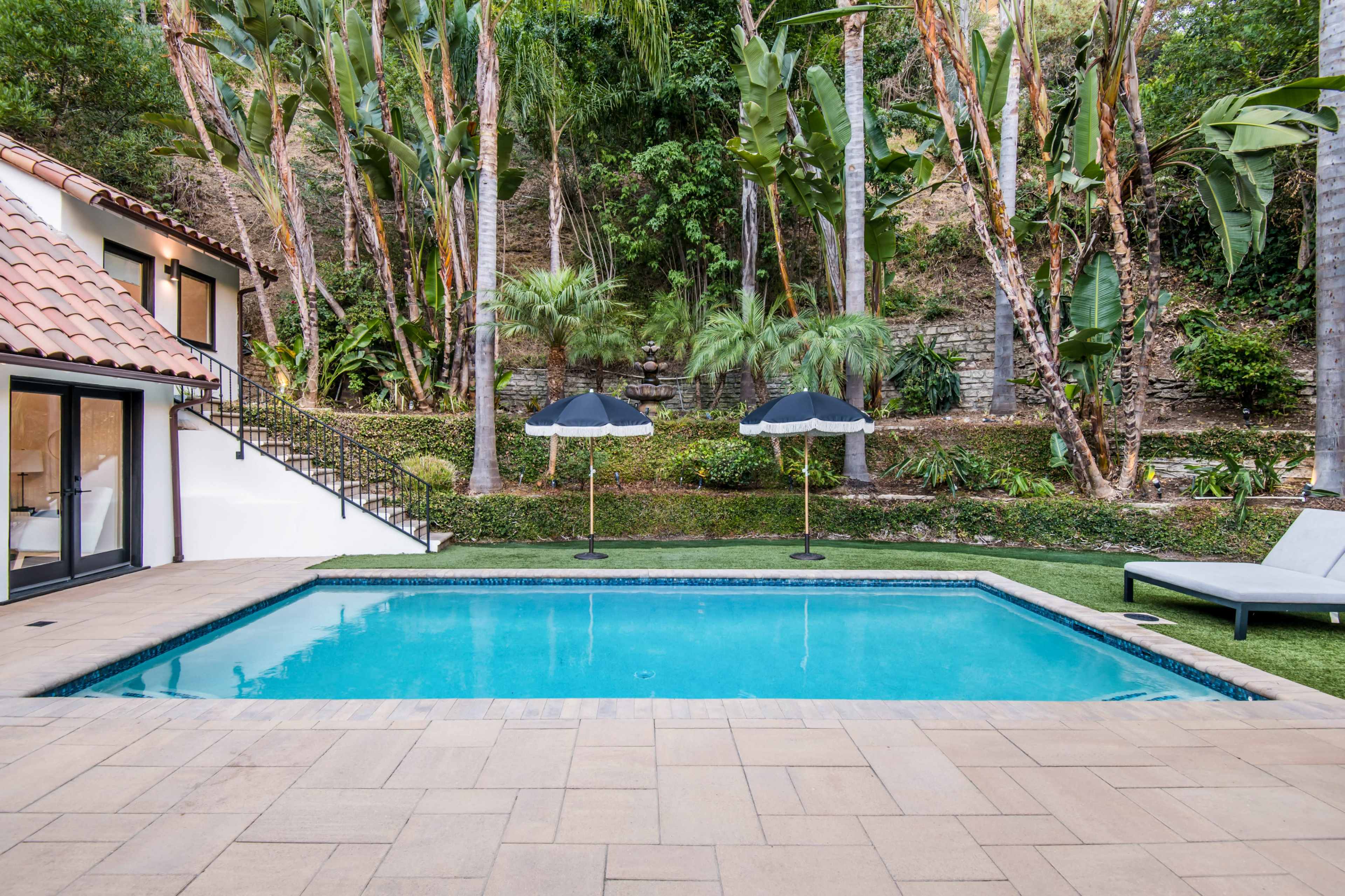 A swimming pool surrounded by greenery, with two umbrellas and a lounge chair nearby.