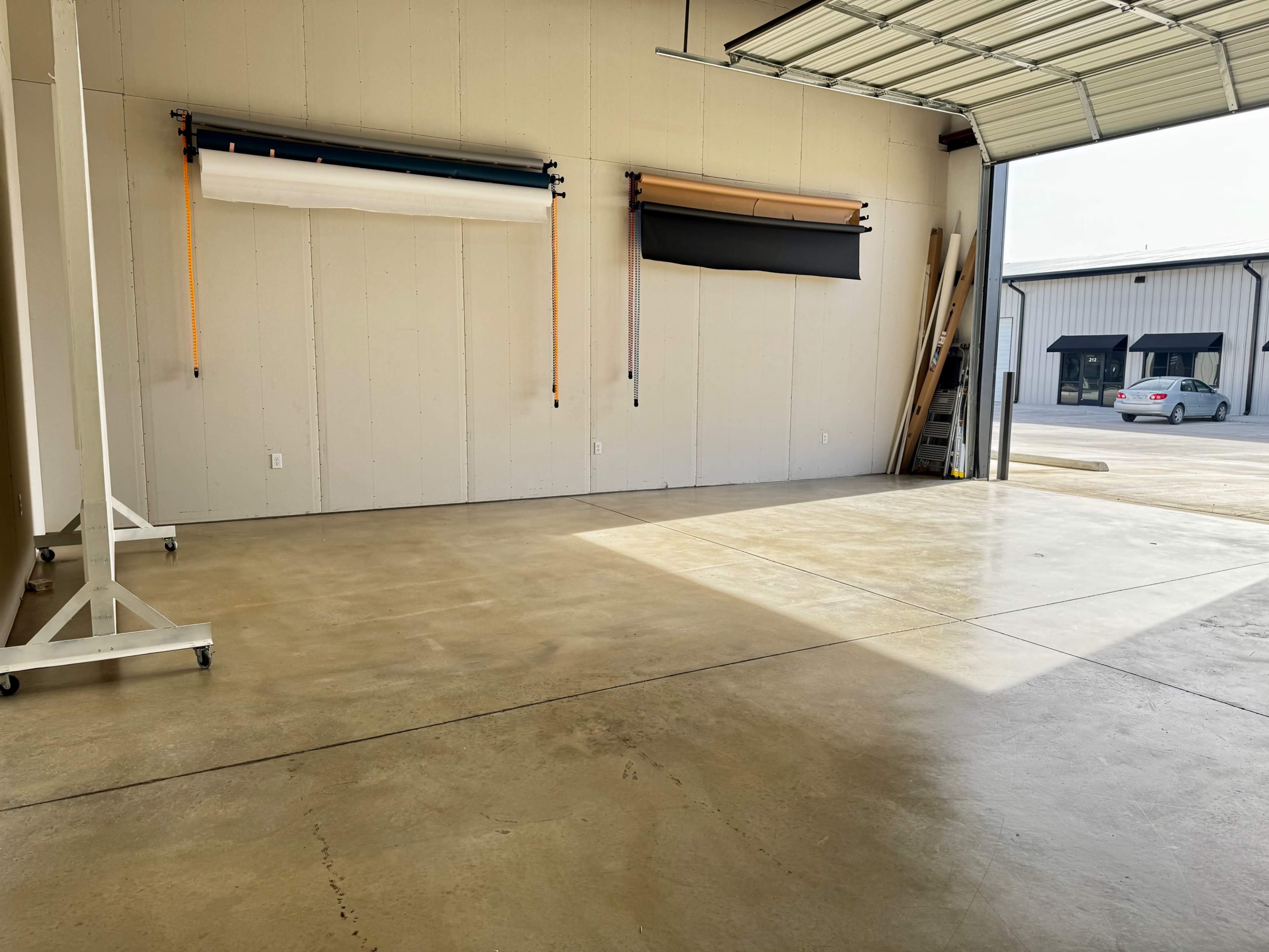 The image shows an empty warehouse space with rolling backdrop material stored on the wall and a vehicle parked outside in the sunlight.