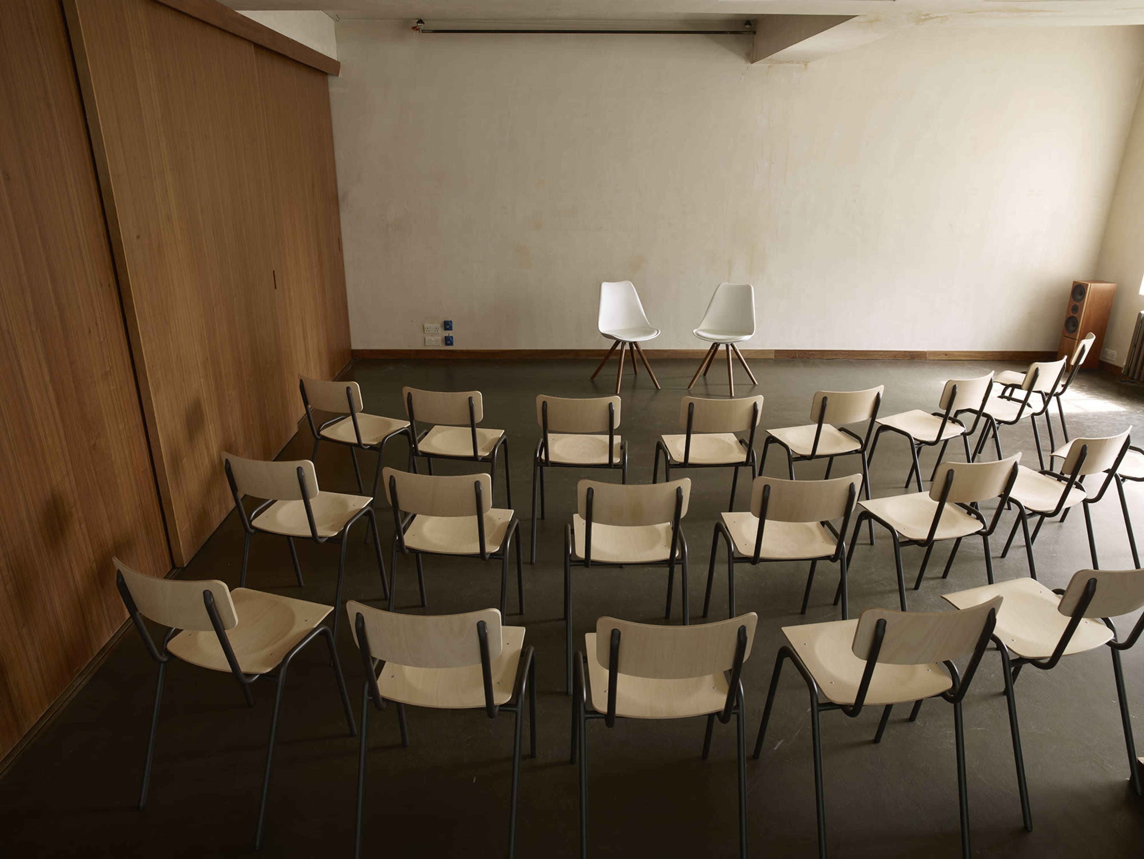 A row of light wooden chairs faces two white chairs on a stage in a sparsely decorated room.