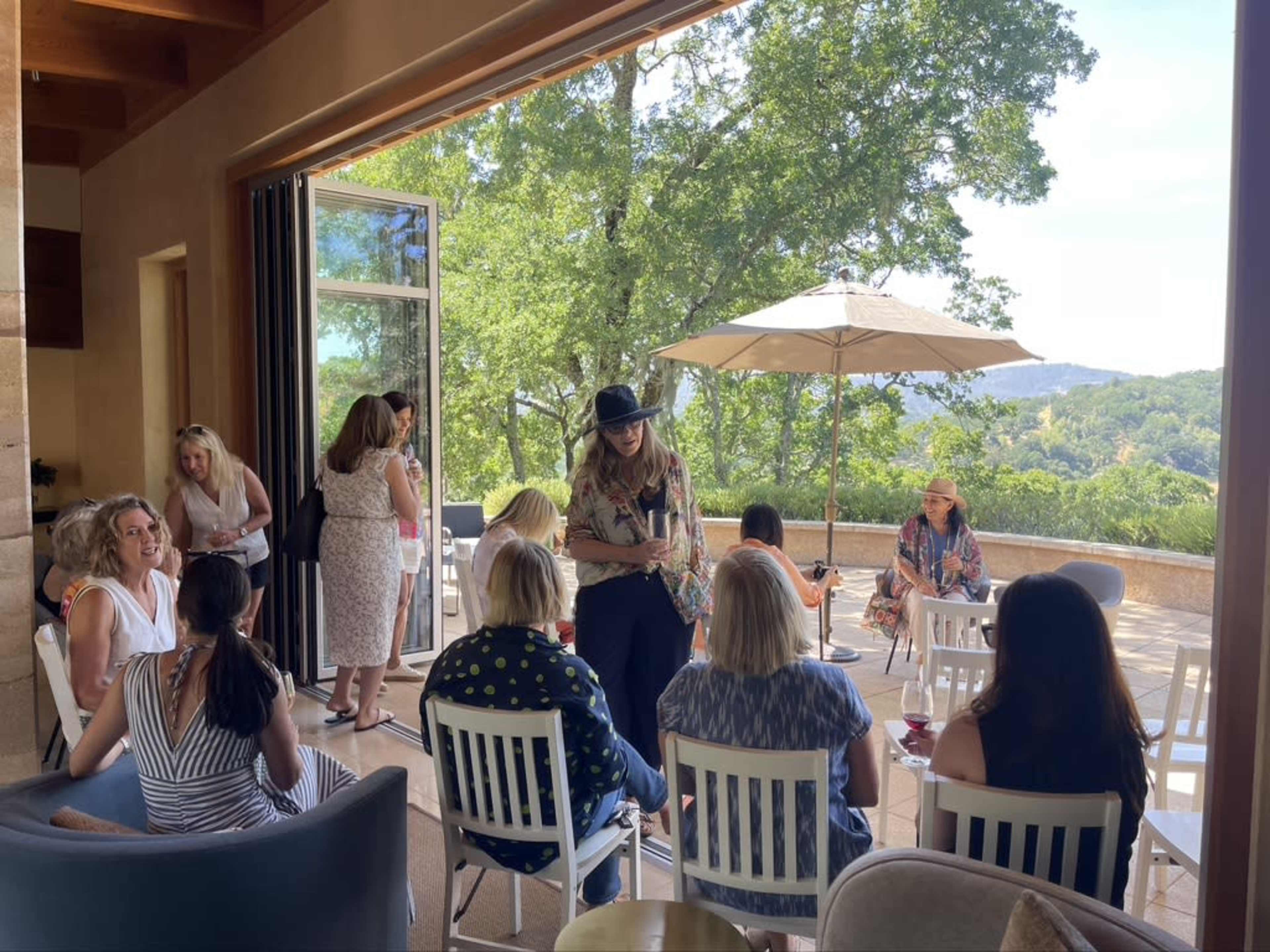 A group of women are gathered in a spacious indoor-outdoor setting, socializing around tables under an umbrella beside a scenic view.