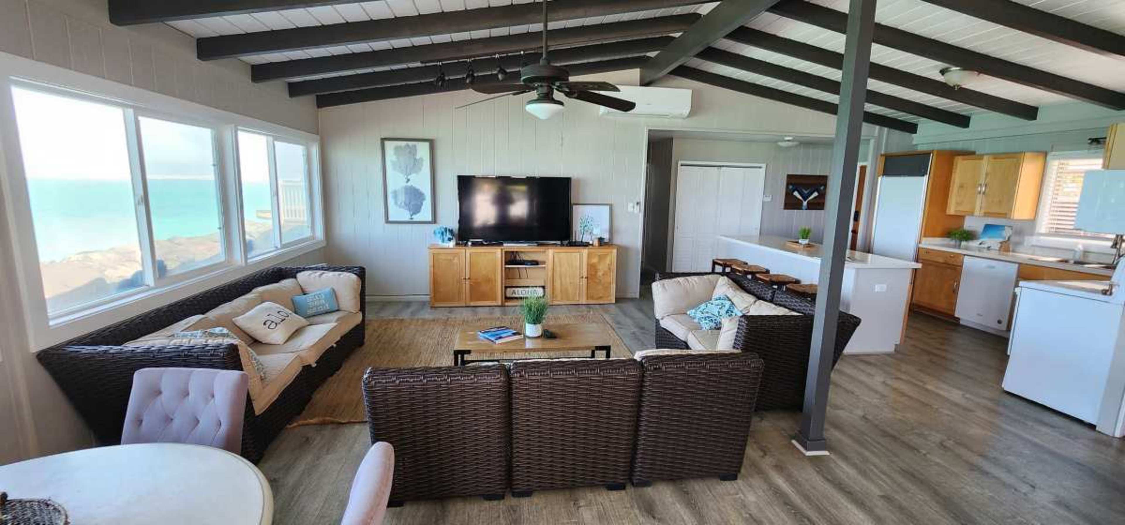 The image shows a modern living space with a seating area, a television, and a kitchen, featuring large windows with a view of the ocean.