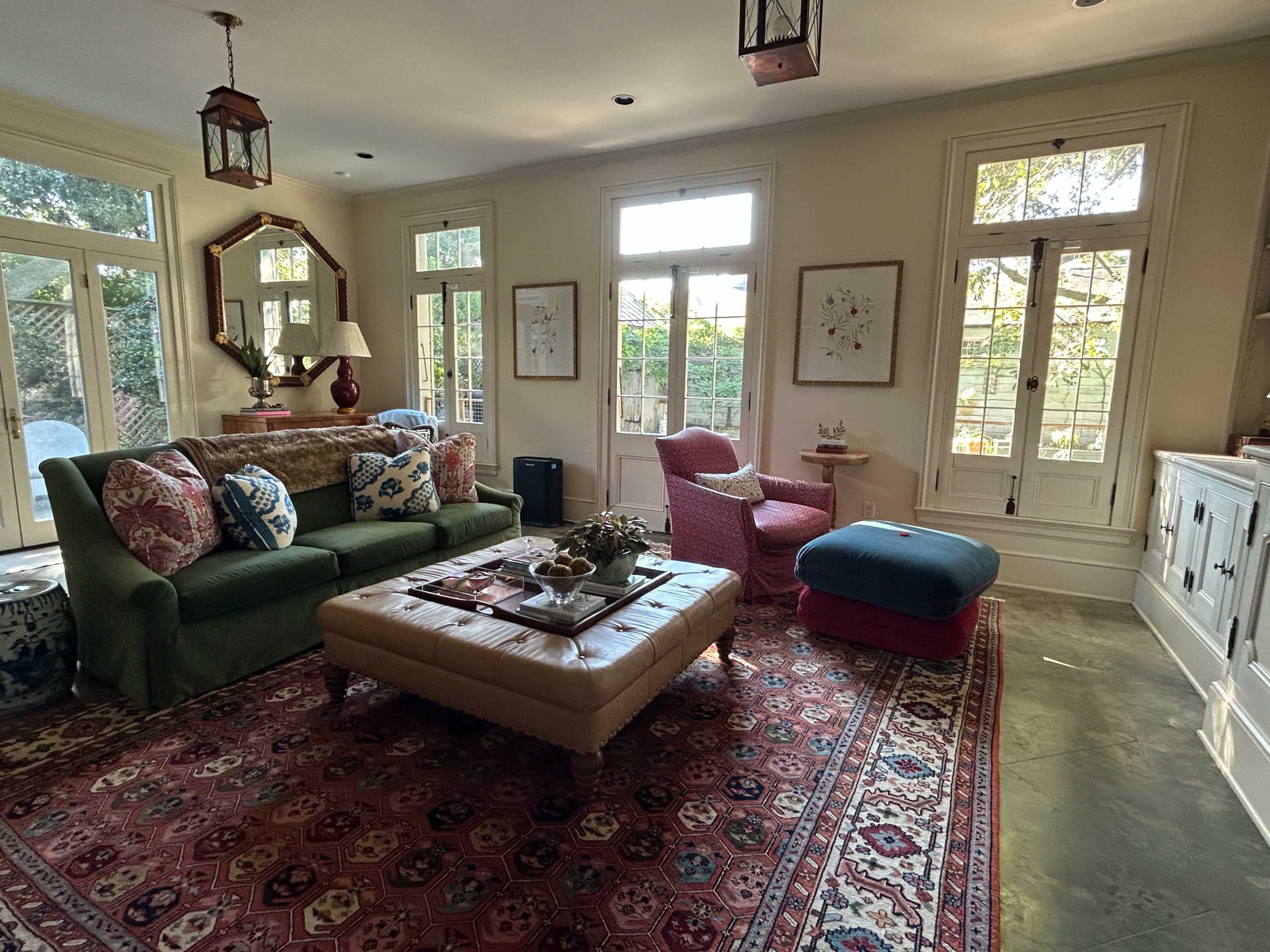 The living room features a combination of green and pink seating, a large ottoman, and windows that let in natural light, surrounded by decorative artwork and plants.