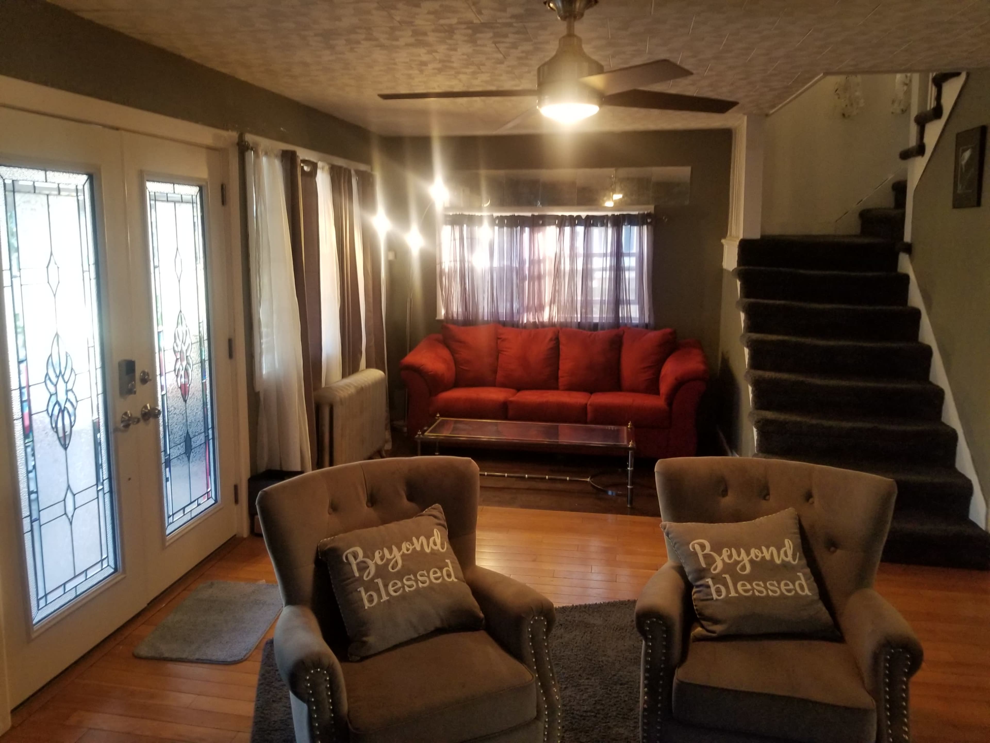 A living room with a red sofa, two gray armchairs with decorative cushions, a wooden coffee table, and a staircase in the background.