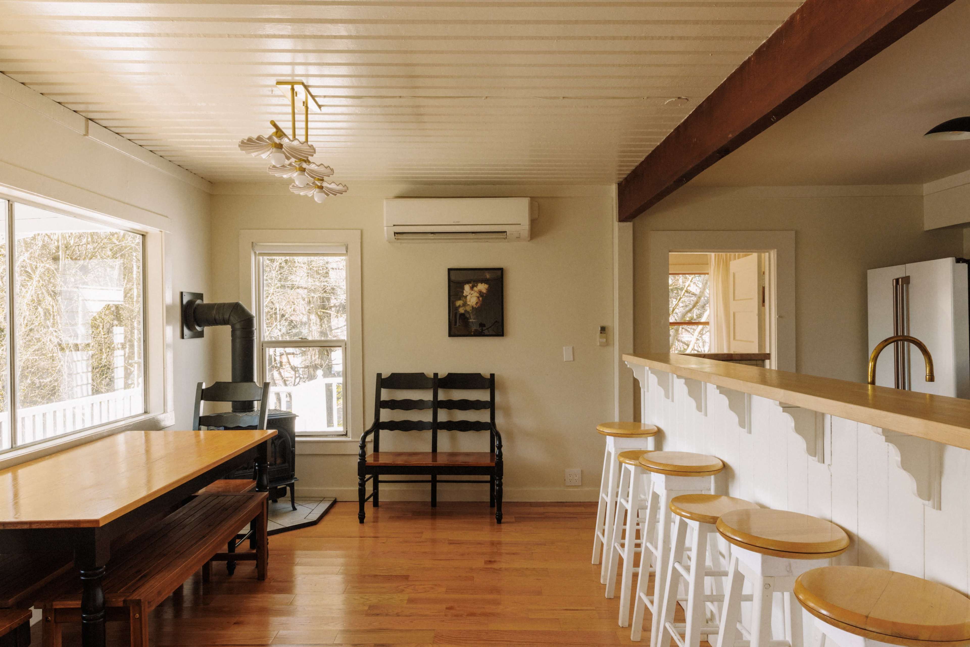 A simple dining area with a wooden table and chairs, a bar with stools, and large windows providing natural light.