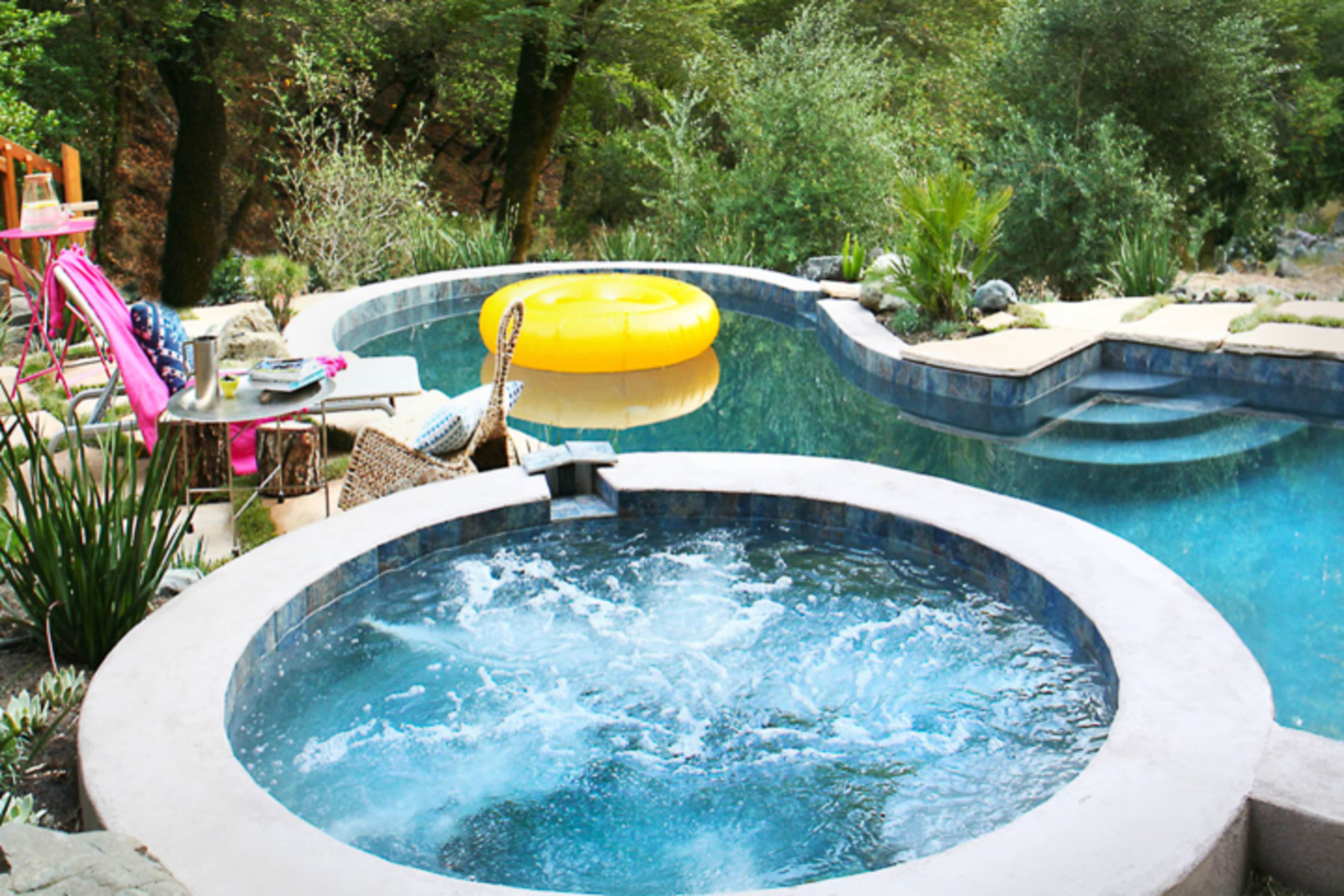 A hot tub bubbles next to a swimming pool surrounded by greenery and a large yellow inflatable float.
