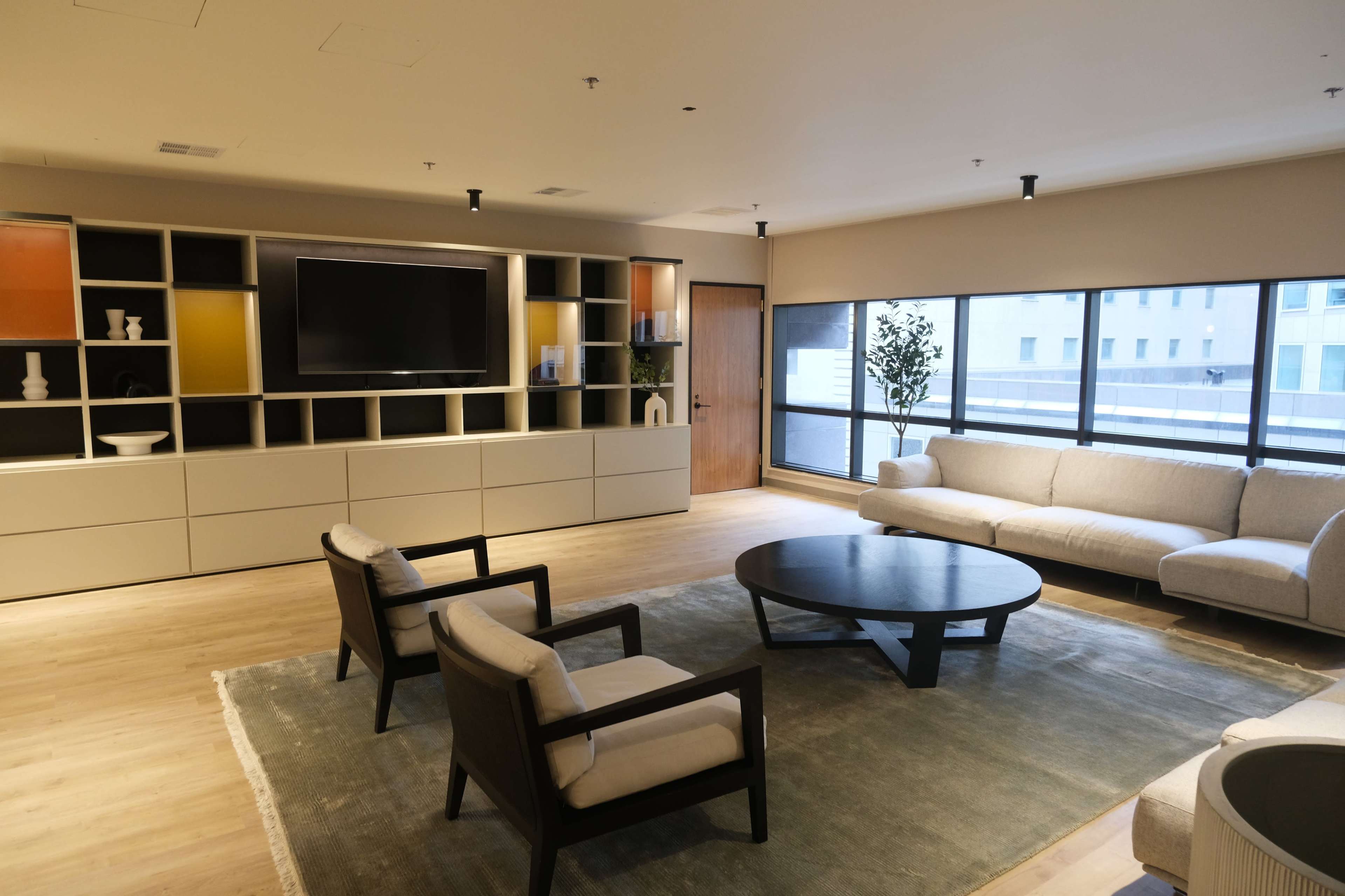 A modern living room with a large TV mounted on a wall unit, a round coffee table, two armchairs, and a sofa facing large windows.