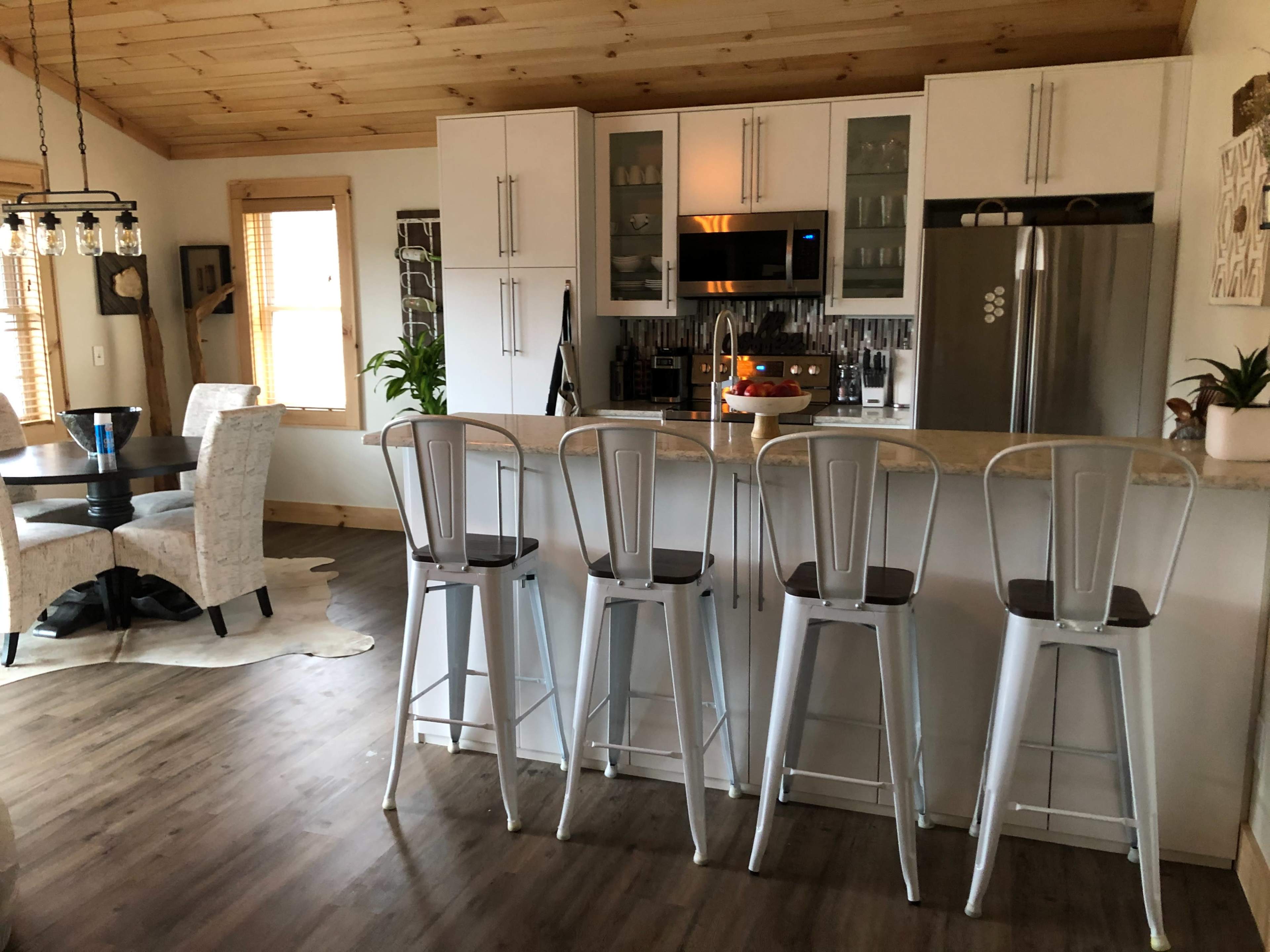 A modern kitchen with a bar area featuring four metal stools, a dining table in the background, and wooden ceiling beams.
