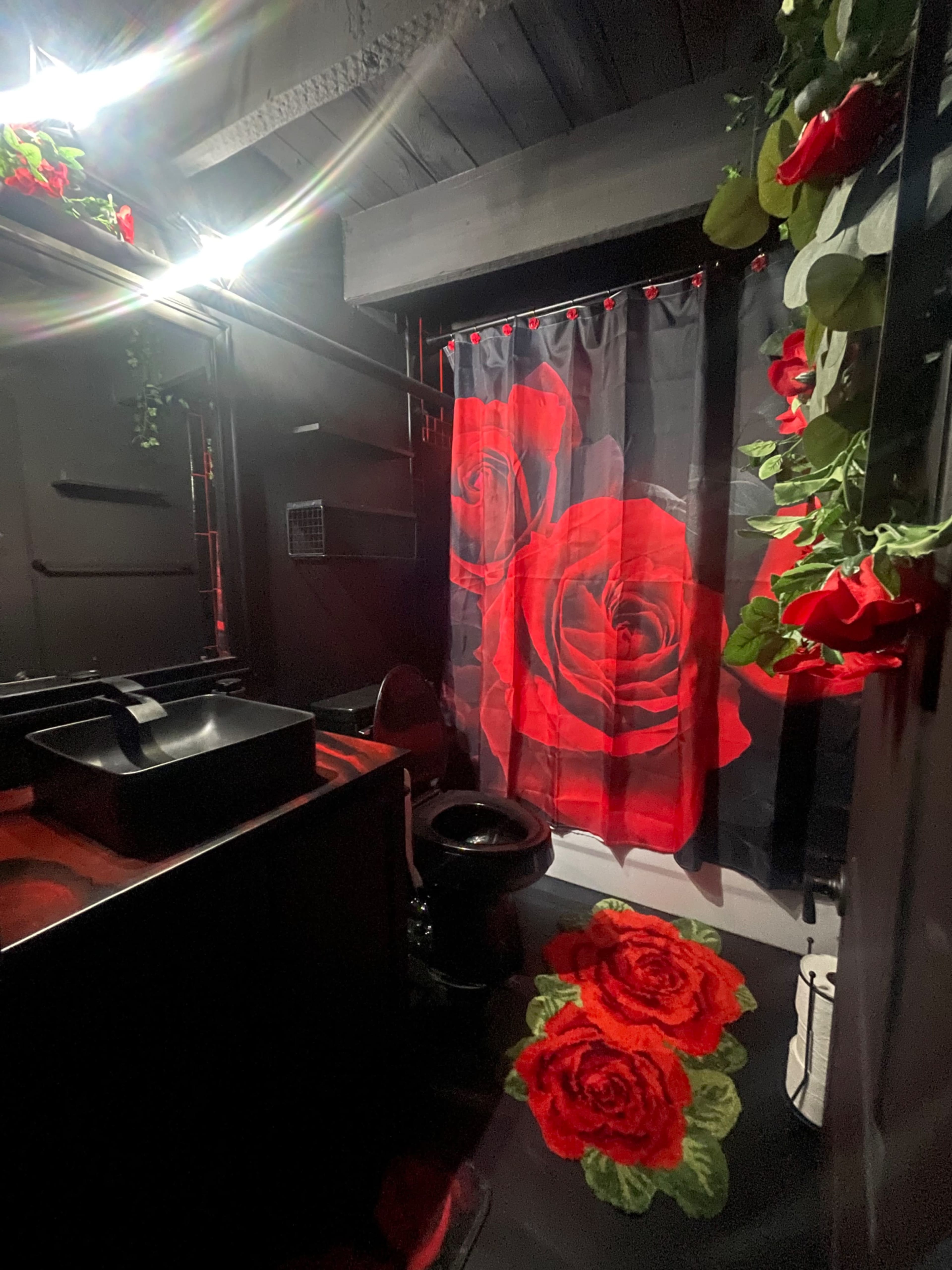 The bathroom features a black and red color scheme, with a shower curtain adorned with large red roses and floral decorations on the walls and floor.
