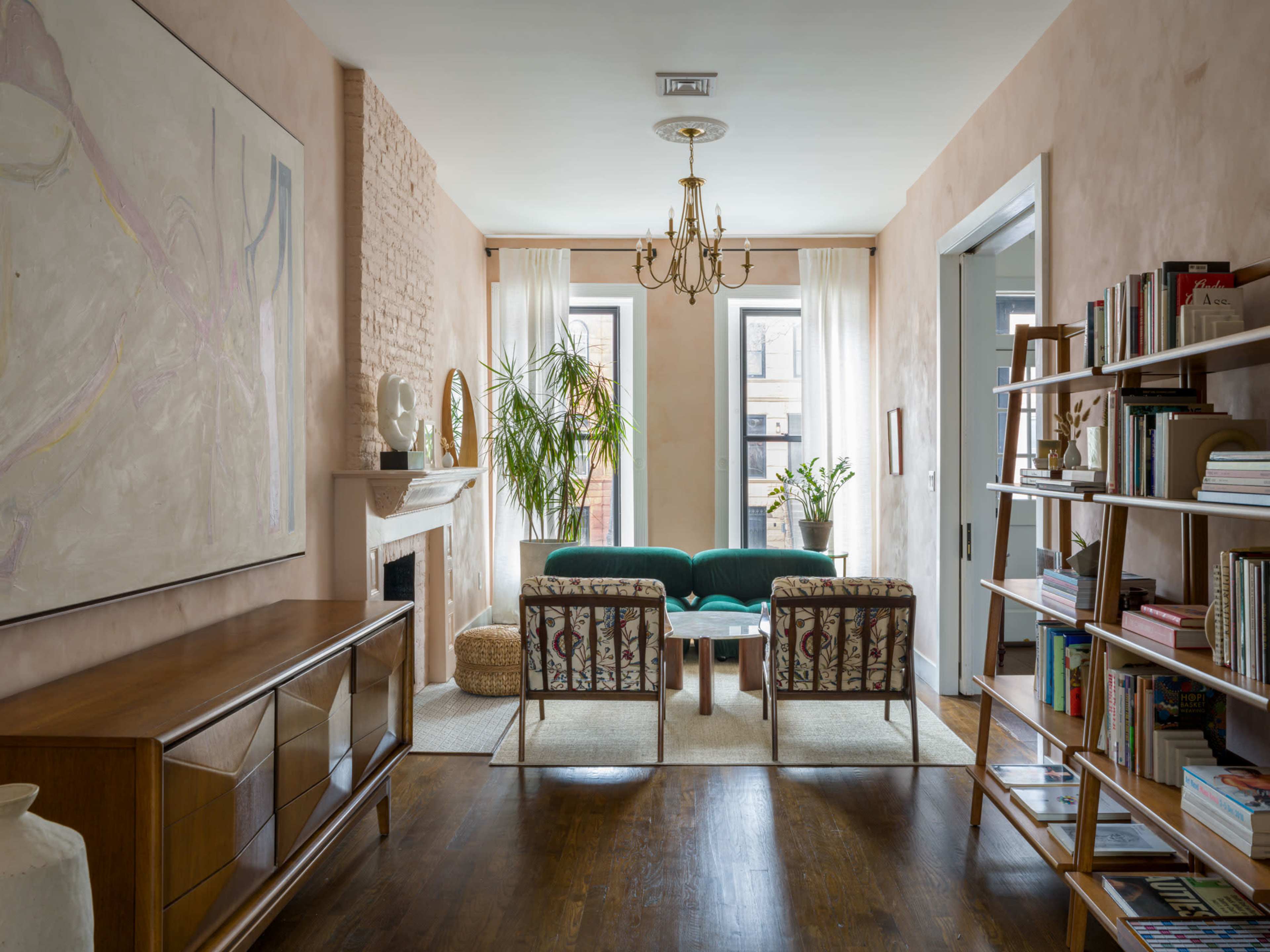 A cozy living room featuring a teal sofa, two patterned chairs, a wooden sideboard, and bookshelves, with large windows allowing natural light to fill the space.