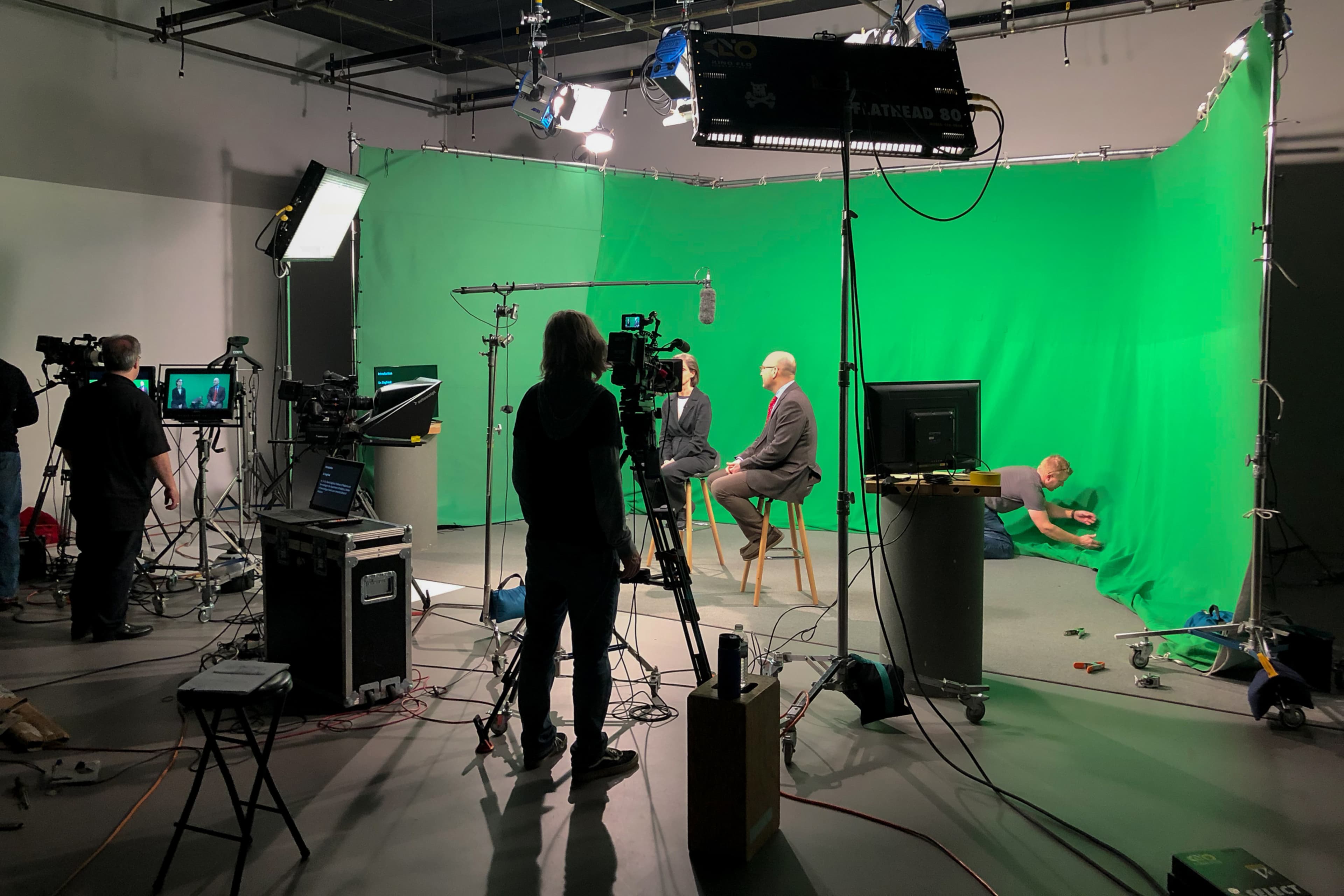 A television production is taking place in a studio with a green screen backdrop, featuring multiple cameras, crew members, and a seated interview setup.