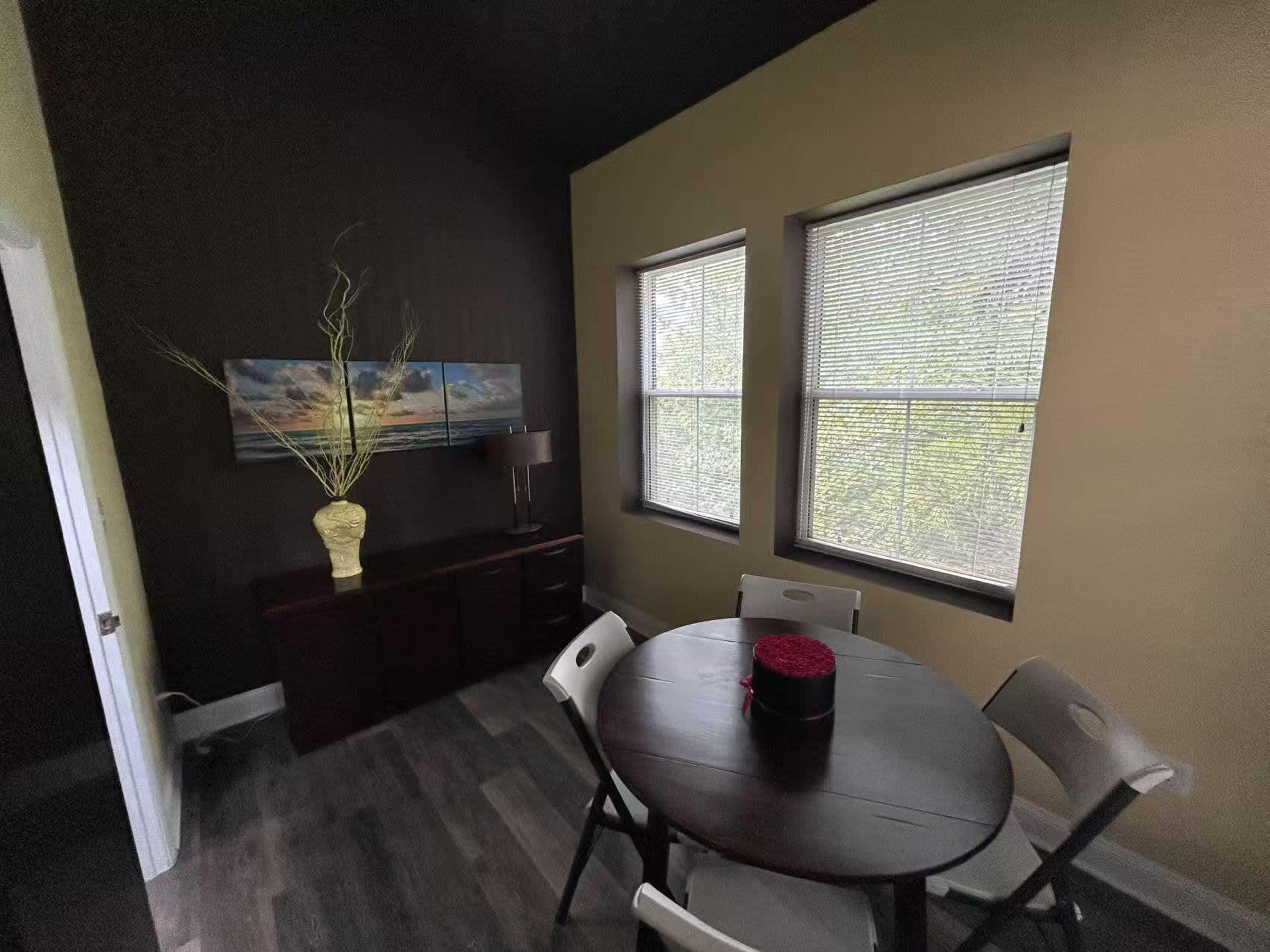 A dining area features a round table with chairs, a decorative vase, and two windows with blinds.