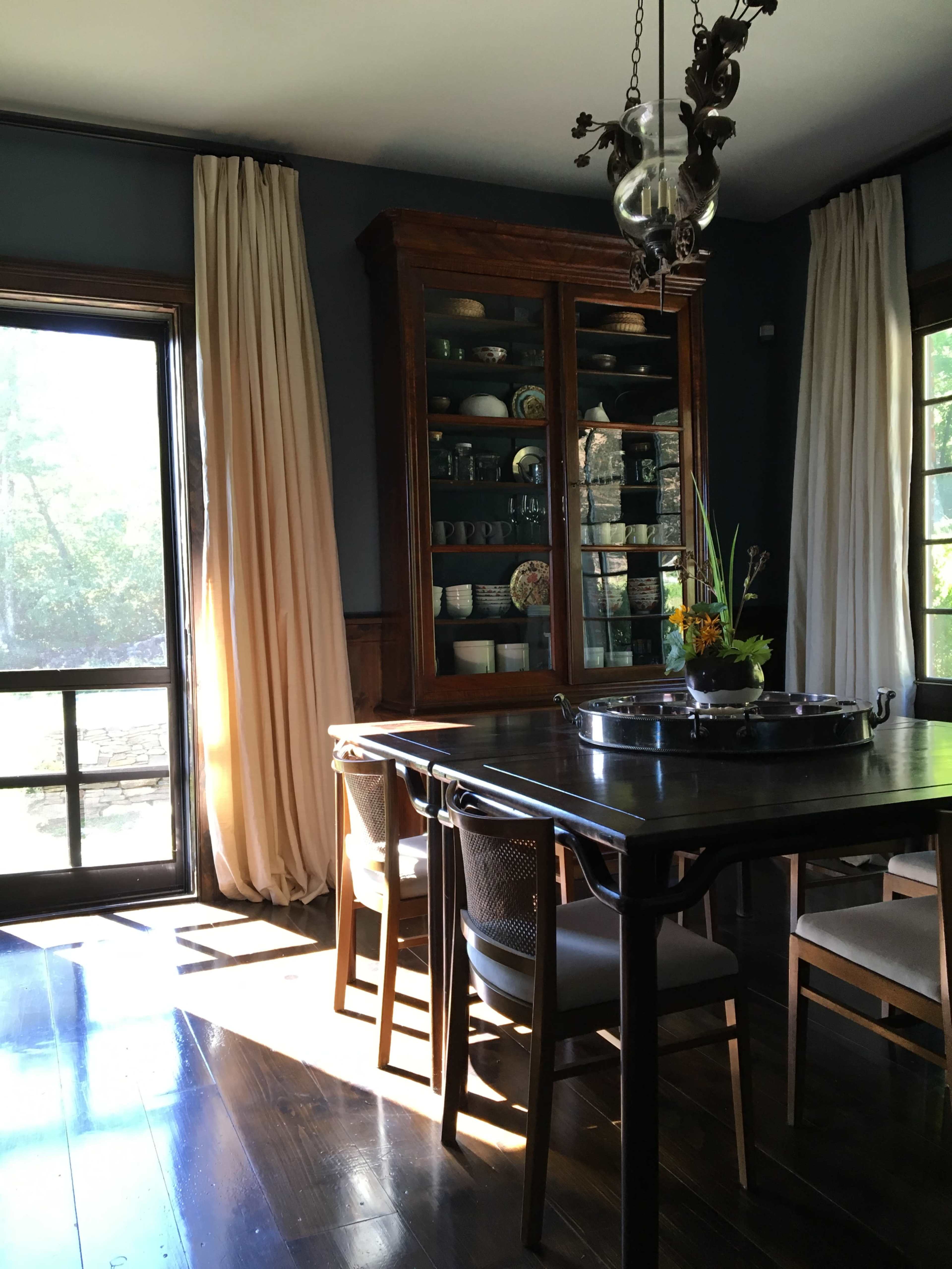 A dining room features a wooden table surrounded by chairs, with a glass-fronted cabinet displaying dishware and natural light coming through a large window.