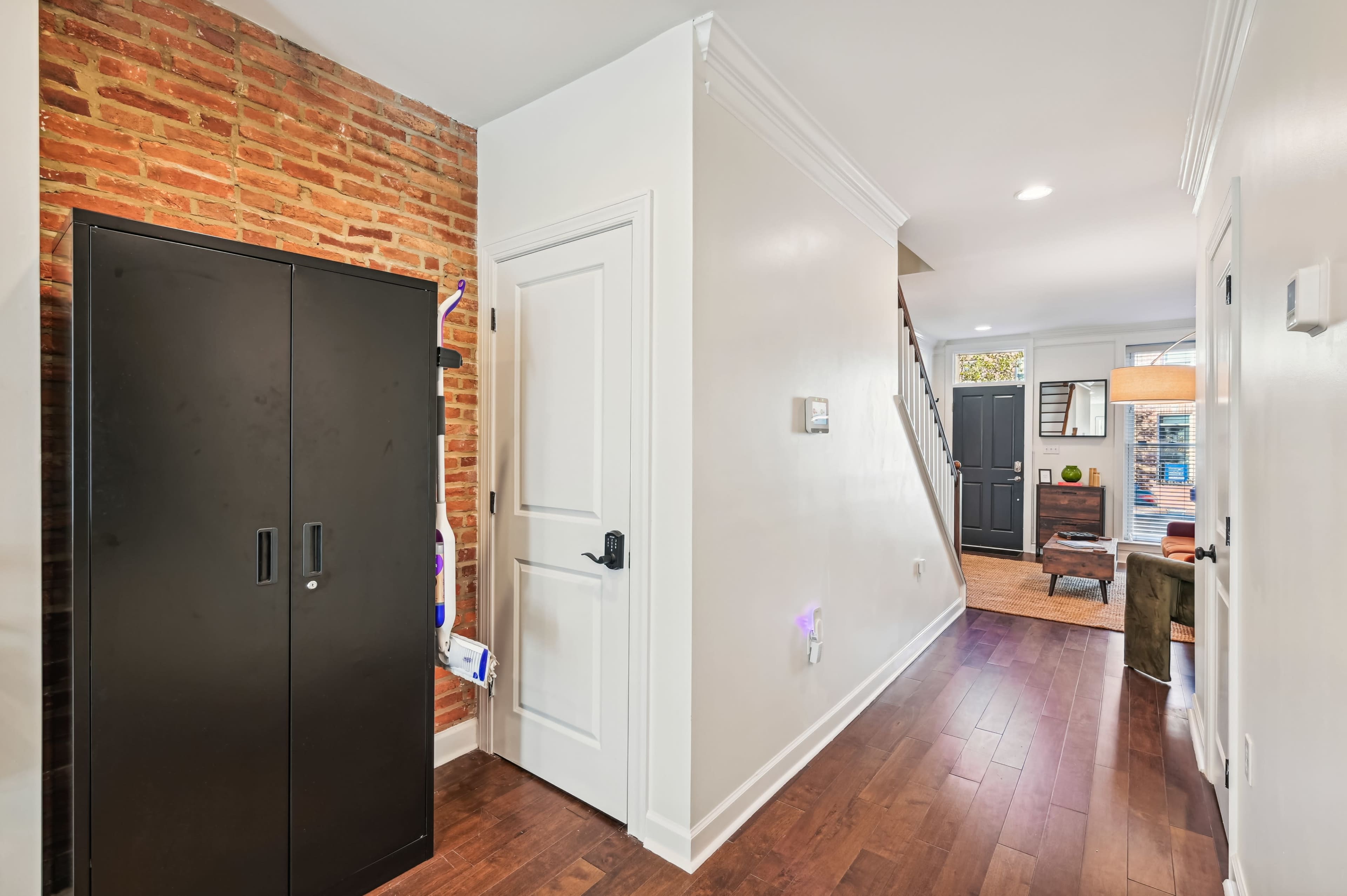 A hallway with a brick accent wall, a black storage cabinet, a closed door, and visible stairs leading to an upper floor.