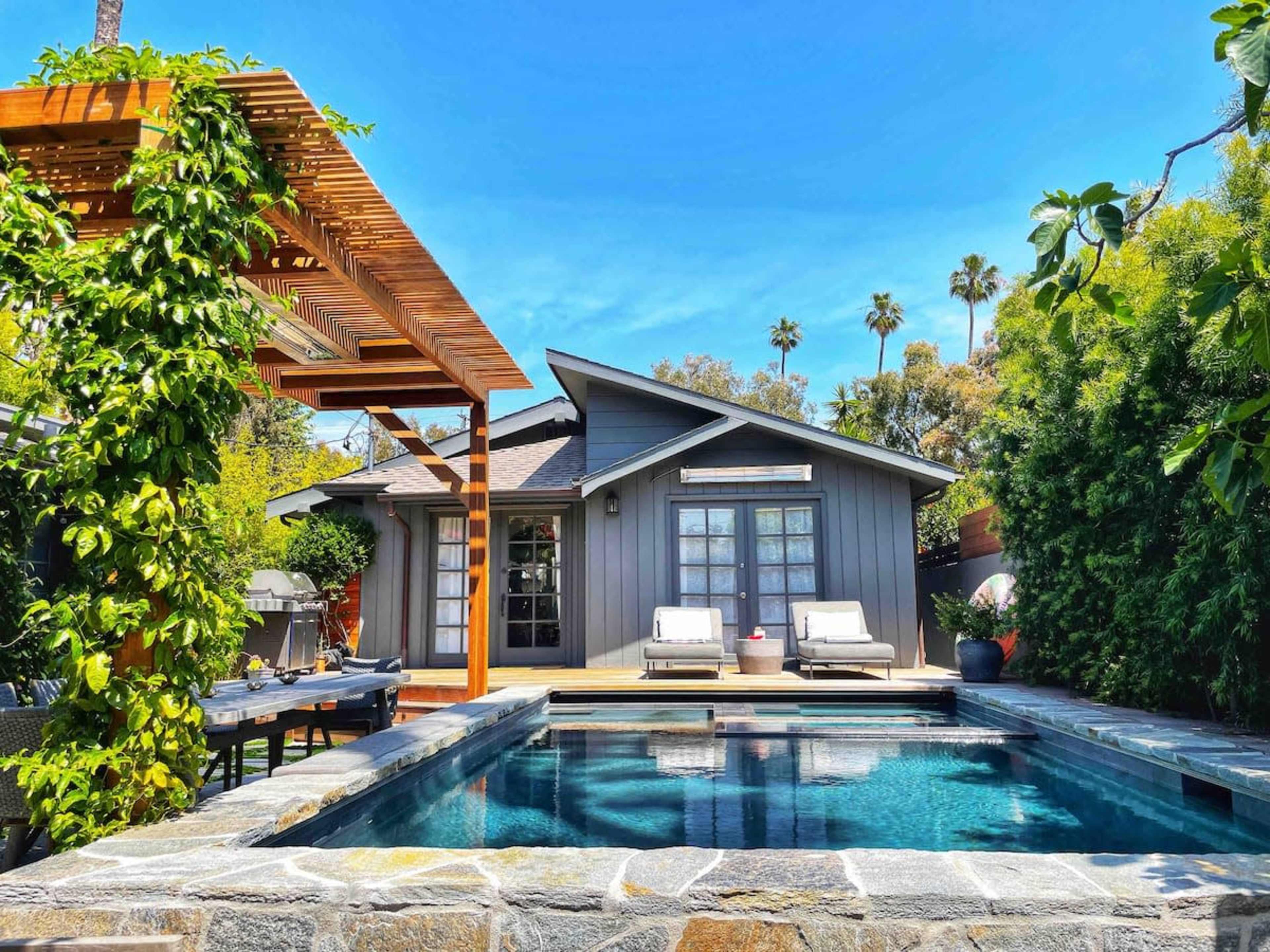 A modern house with a gray exterior sits beside a swimming pool surrounded by greenery and patio furniture.