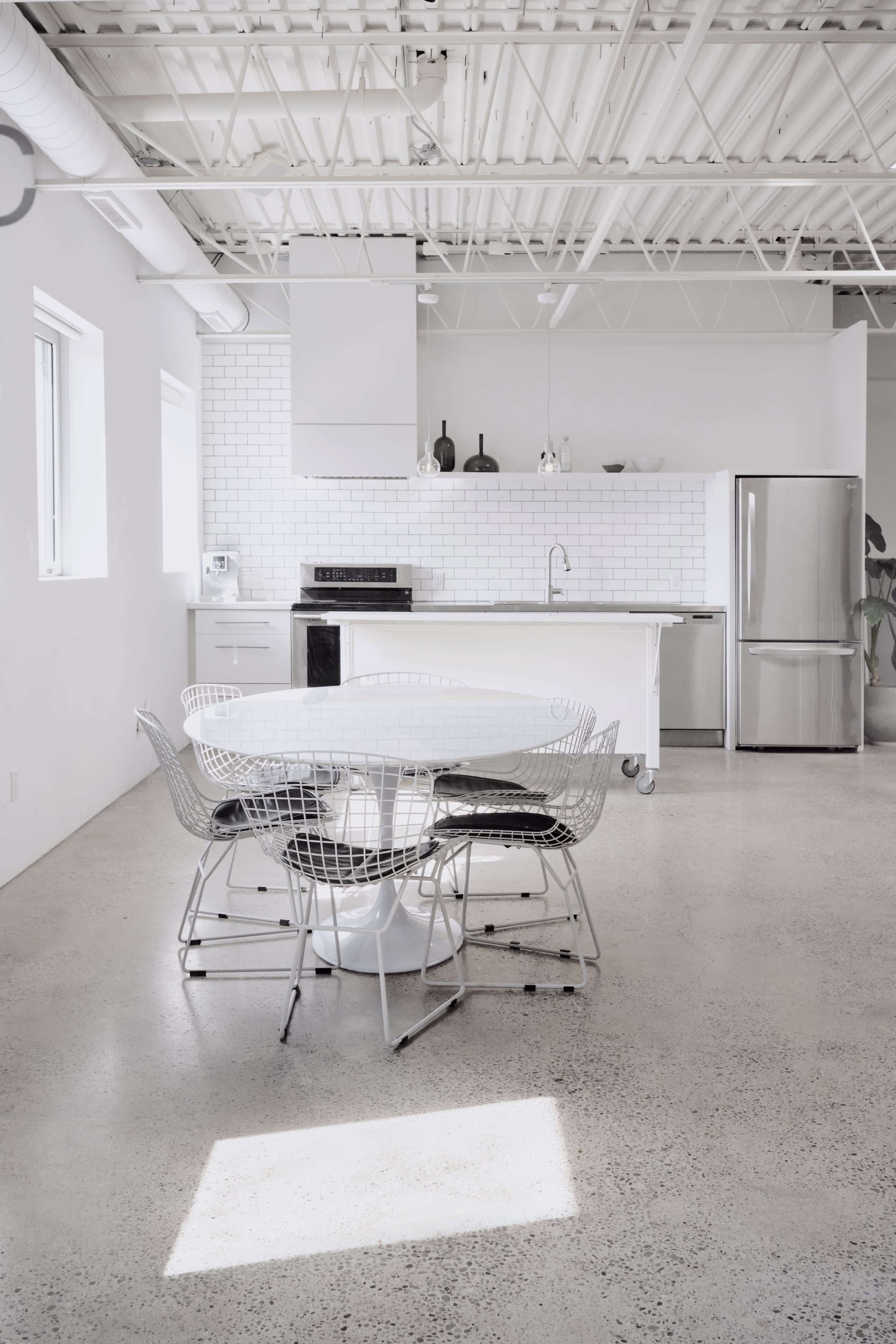 The image shows a modern kitchen and dining area with a round table surrounded by wire chairs, and a kitchen featuring white cabinetry and stainless steel appliances.