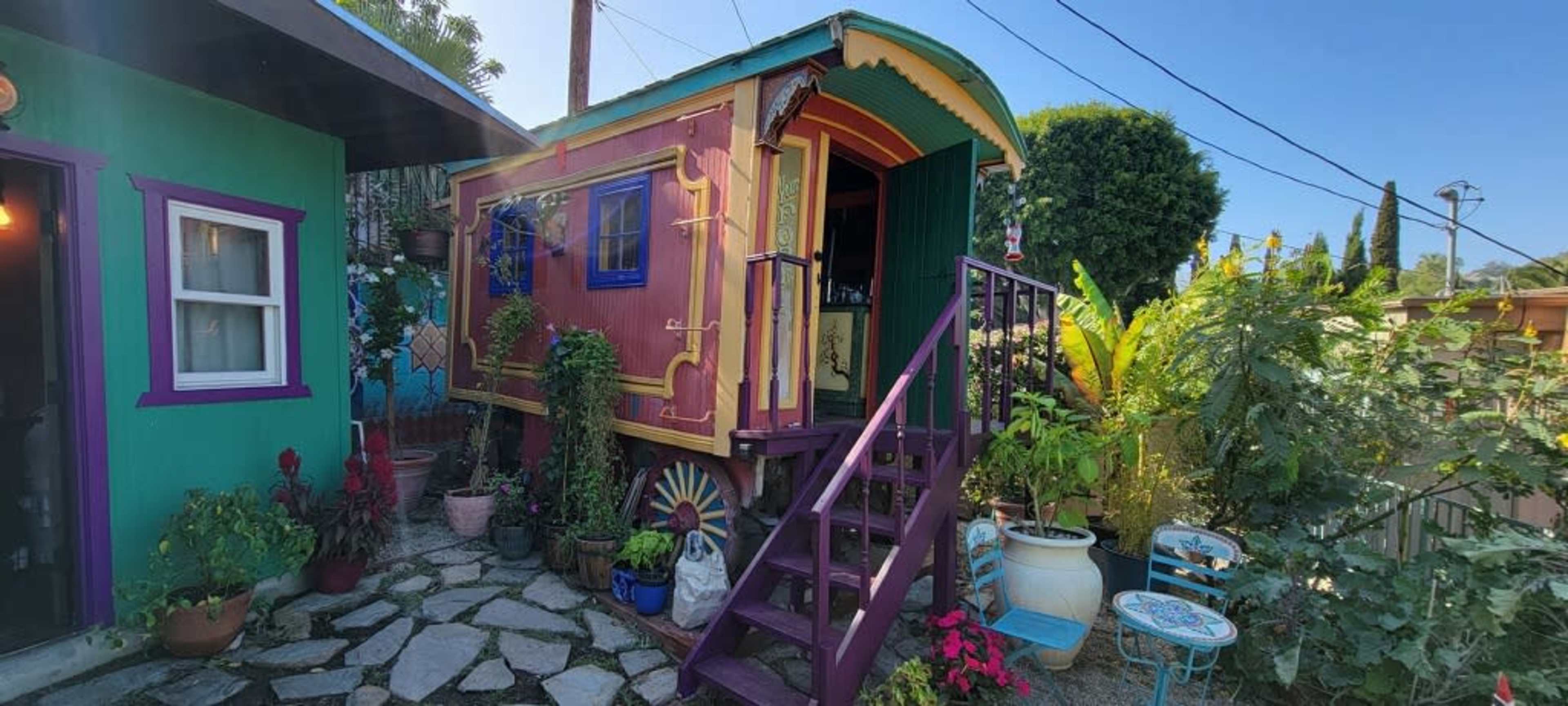 A colorful caravan with a decorated exterior is positioned on a stone path surrounded by various plants and pots.