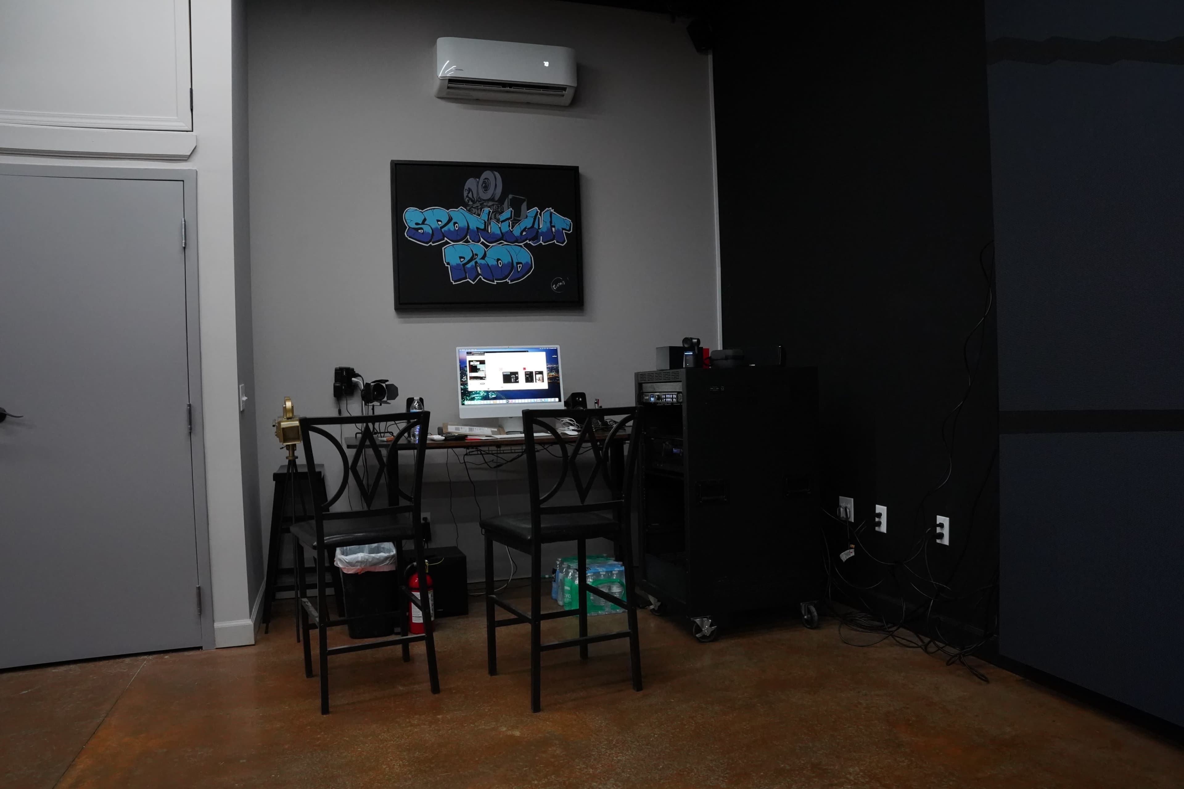 A workspace with two black stools and a desk holding a computer, surrounded by various equipment and a wall-mounted air conditioning unit.