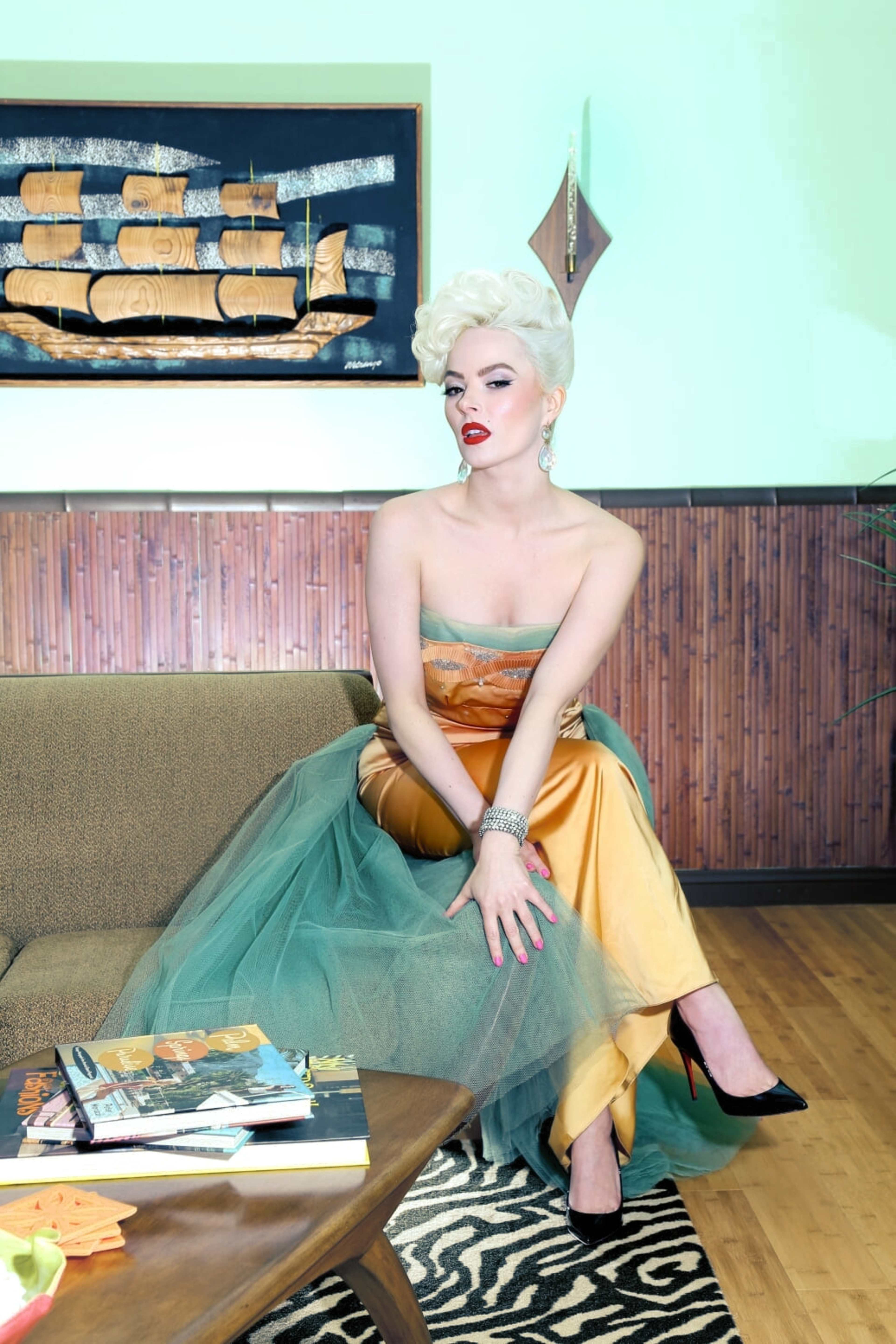 A woman in an elegant gown with flowing fabric sits on a mid-century modern couch, surrounded by decorative items in a stylish room.