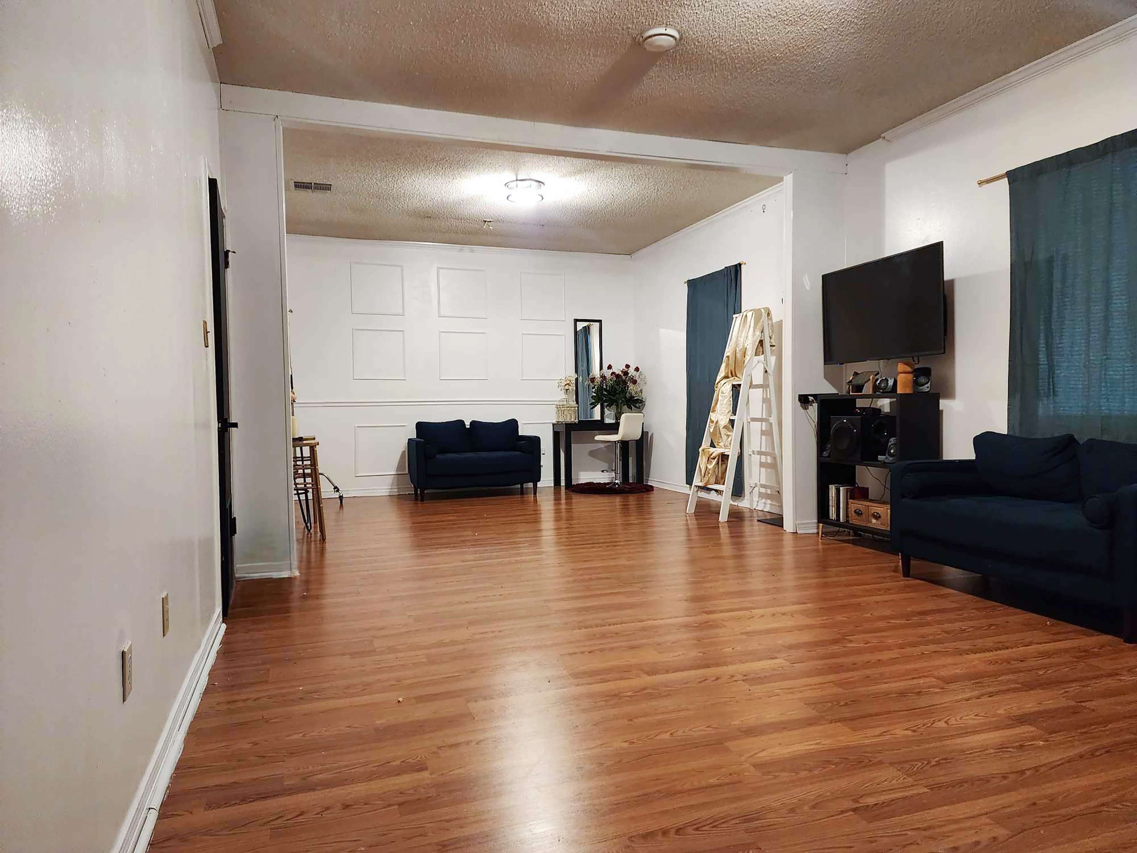 A spacious, well-lit room featuring two navy blue couches, a television, a tall ladder shelf, and a small dining area in the background.