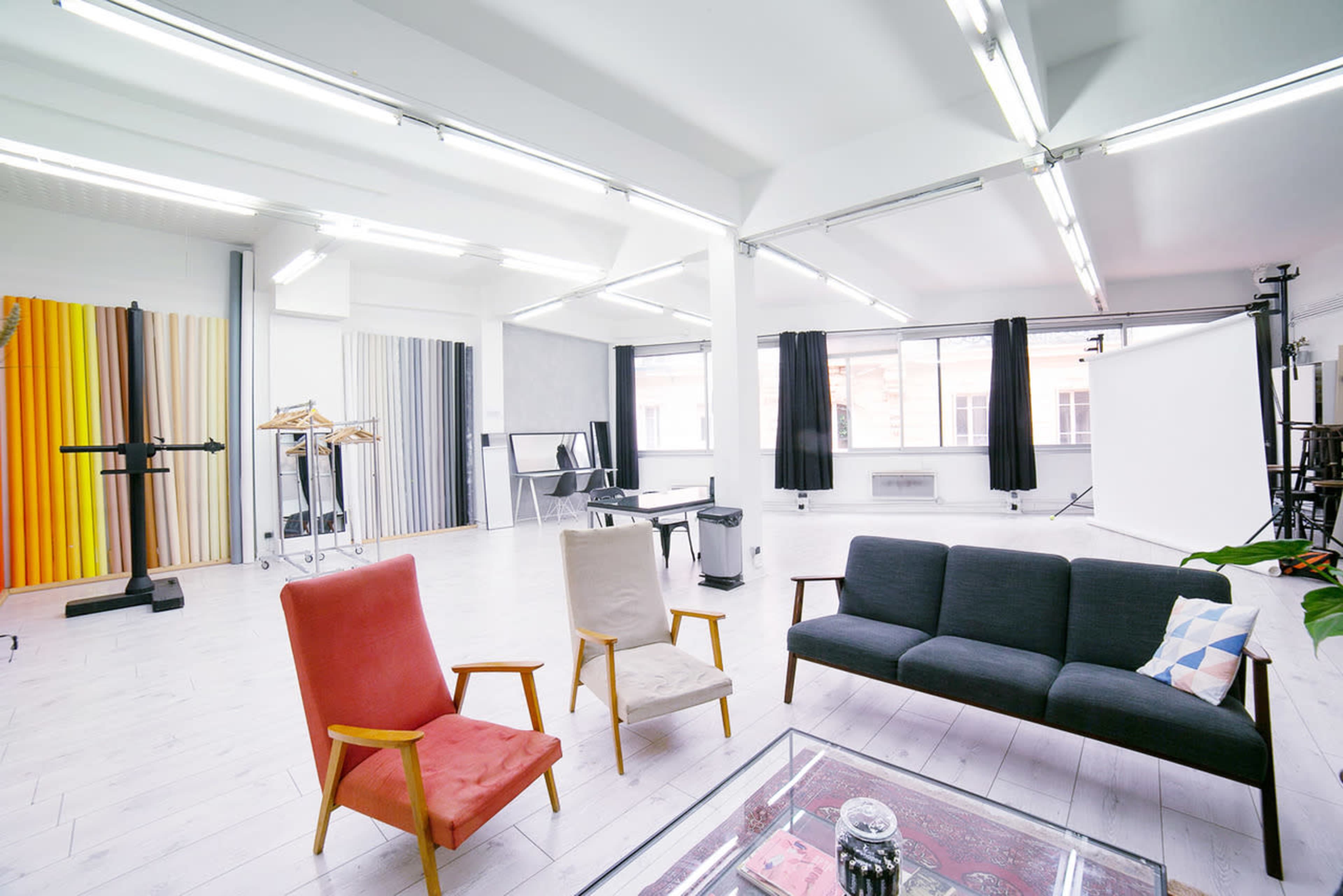 A bright, spacious studio featuring a sofa, two armchairs, and several backdrop stands against a large window.