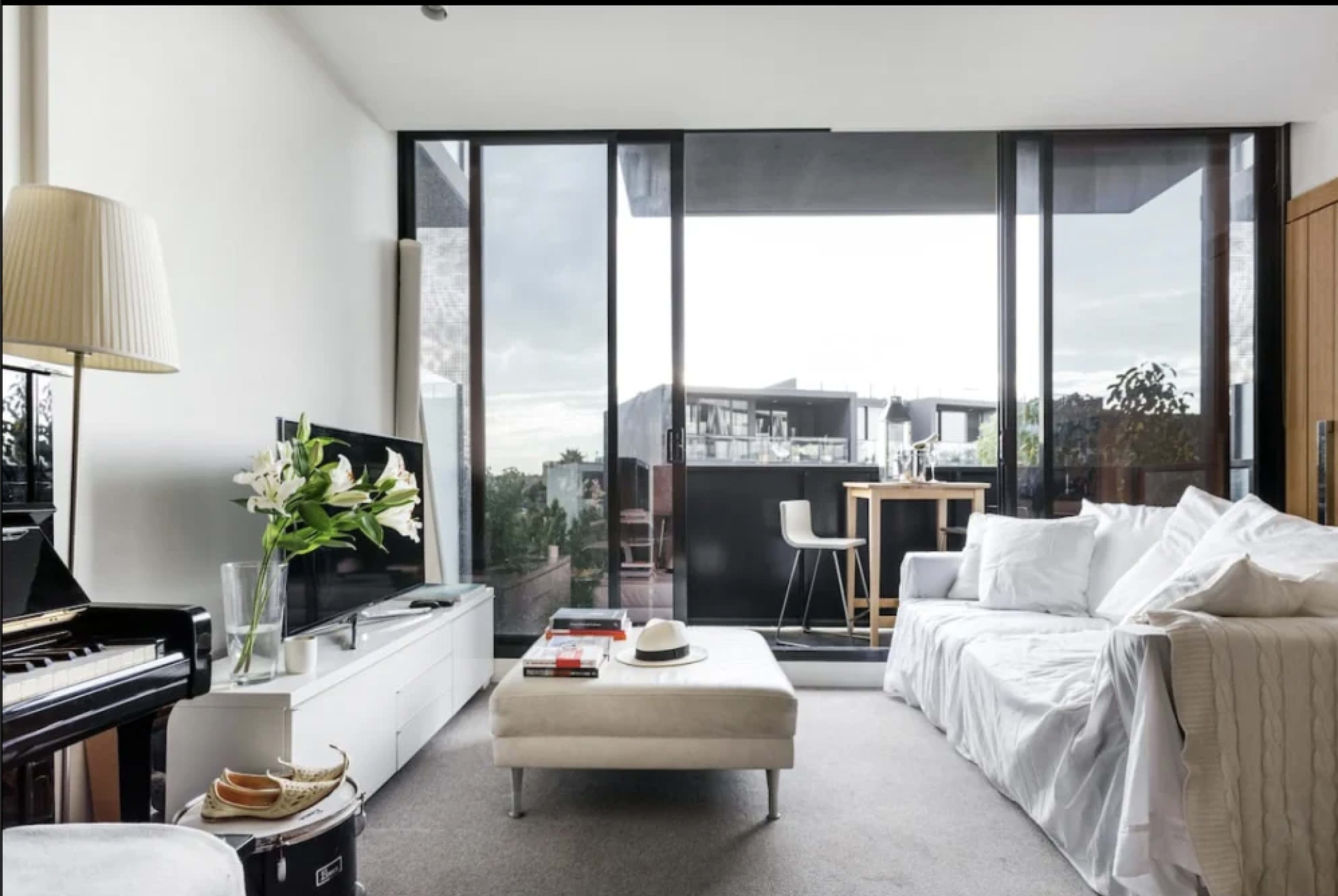 The living room features large windows overlooking a balcony, a white sofa, a piano, and a minimalist coffee table.