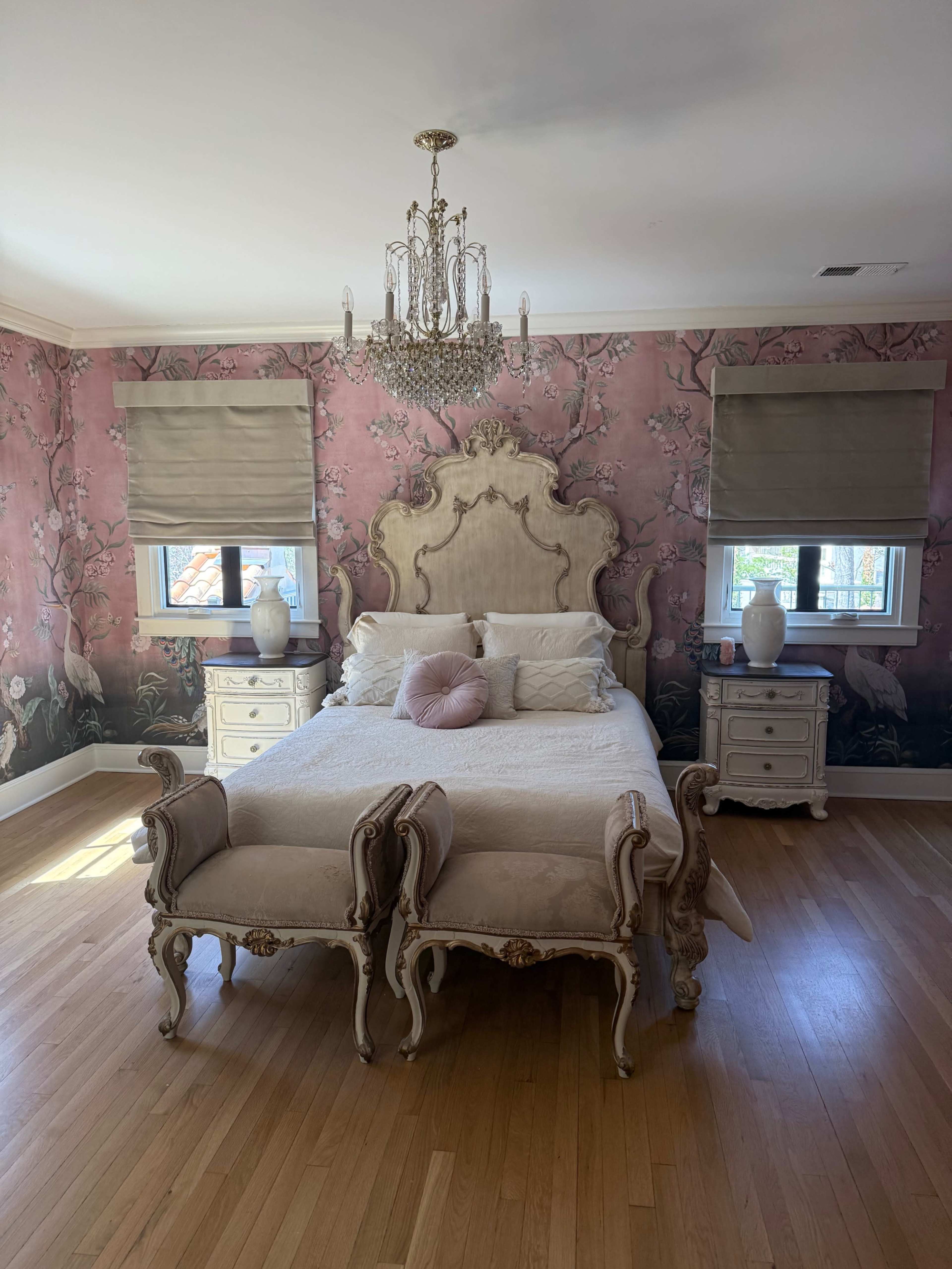 The bedroom features a vintage-style bed with an ornate headboard, flanked by two nightstands, against a floral wallpaper and adorned with a chandelier.