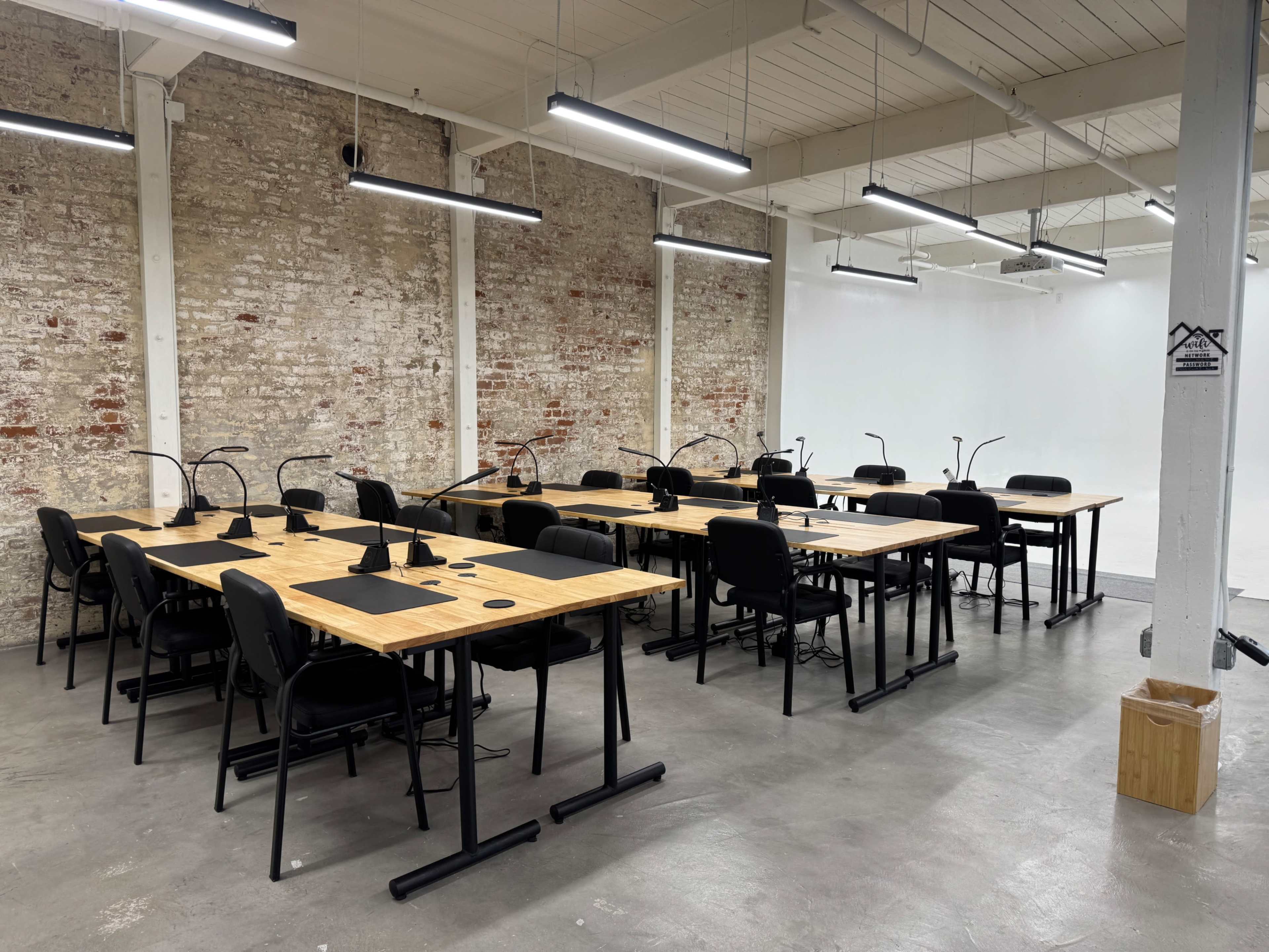 The image shows a spacious, modern coworking area featuring several wooden work tables, black chairs, and task lighting, set against a backdrop of exposed brick walls.