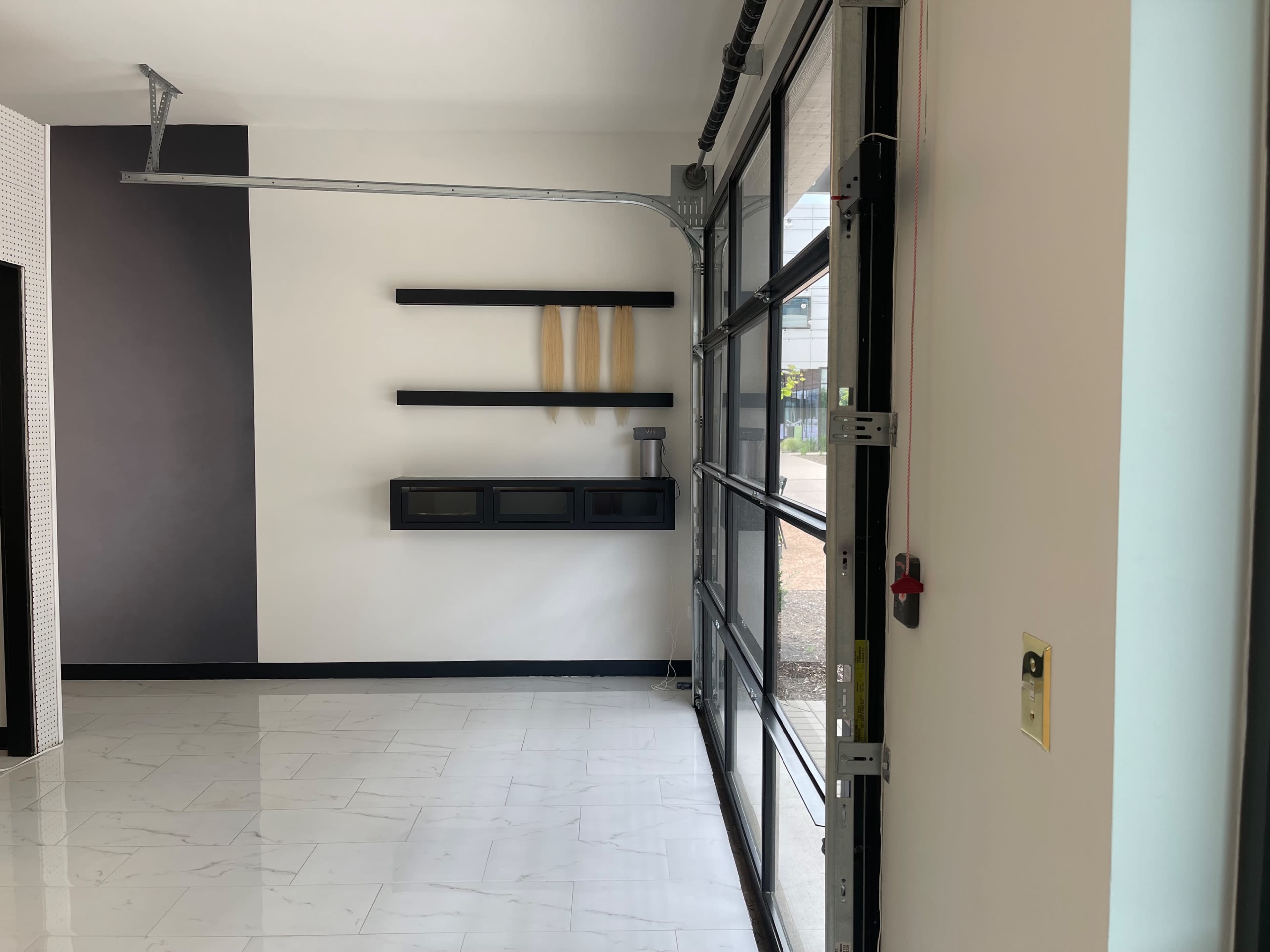 The image shows a spacious interior with a large glass door on one side, white walls, a black shelf holding three items, and a light-colored tiled floor.