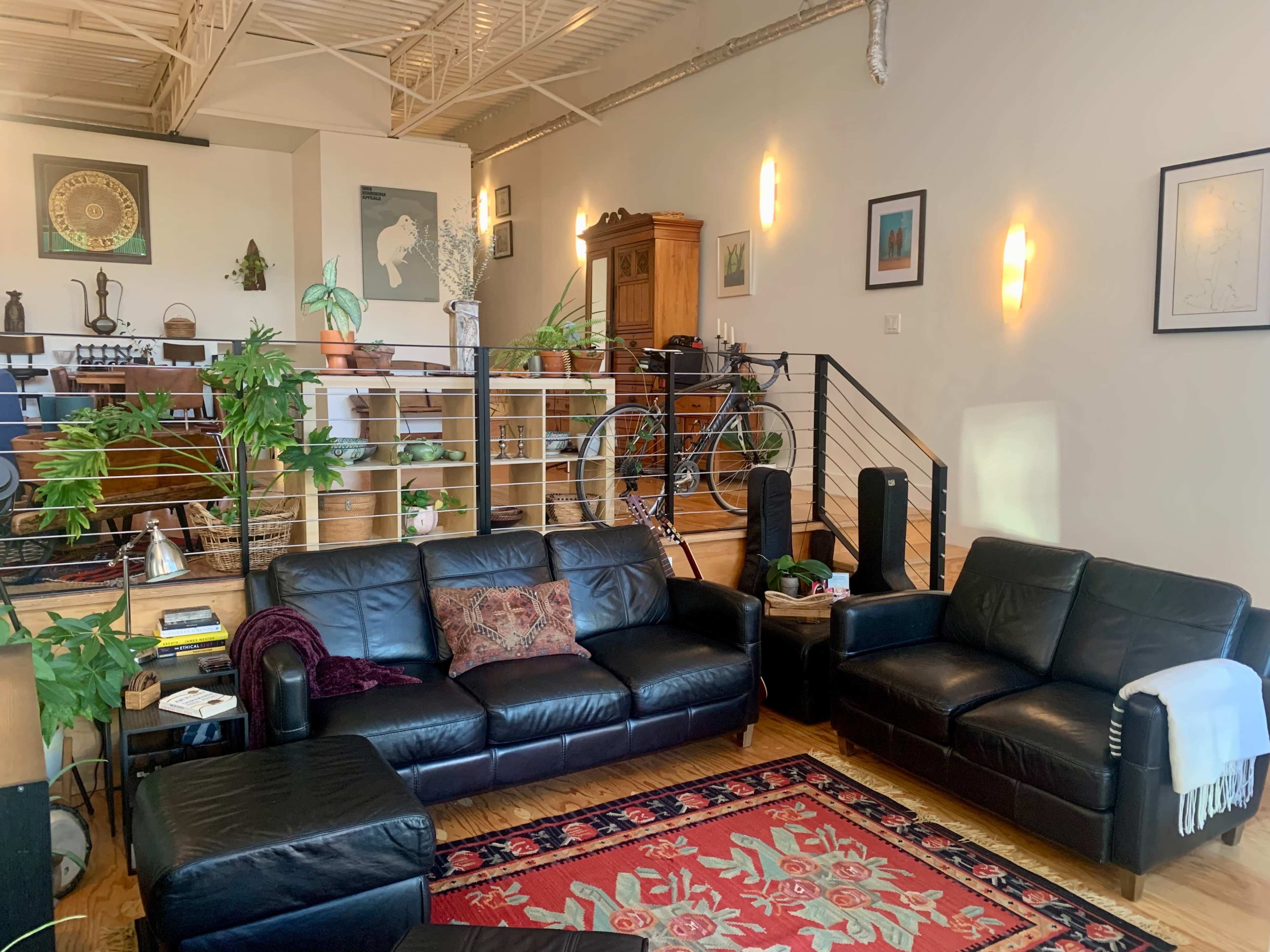 A modern living room with black leather sofas, a coffee table, and a variety of plants, leading to an elevated dining area with wooden furniture and a bicycle in the background.