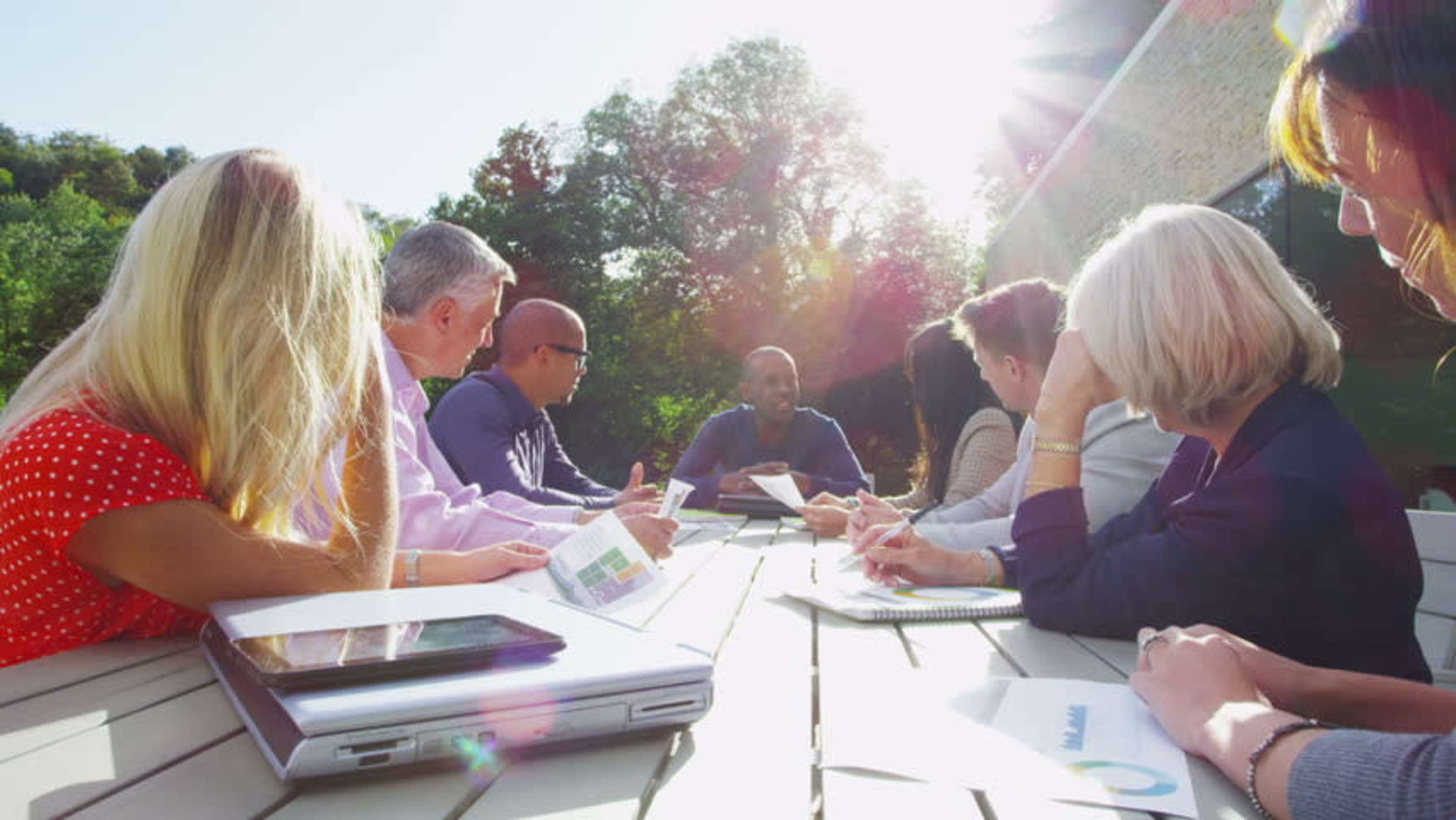A diverse group of professionals is seated around a table outdoors, engaging in a discussion while reviewing documents.