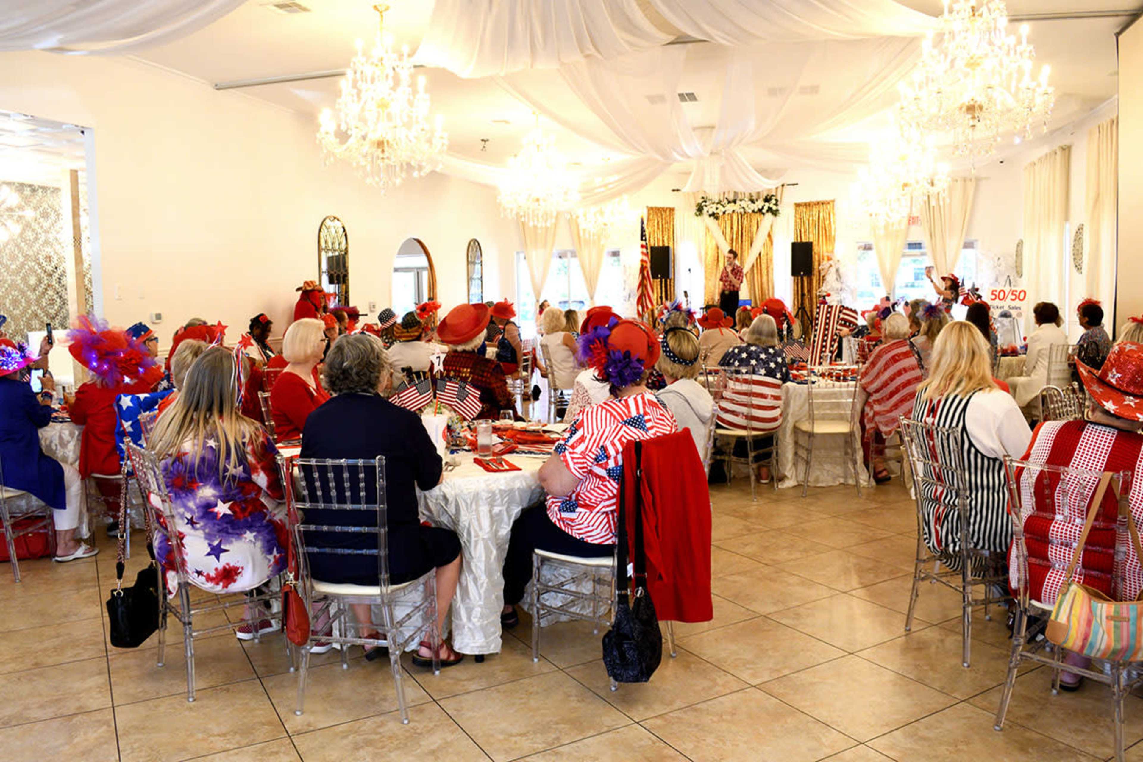 A large group of people in festive attire are seated around tables in a banquet hall decorated with chandeliers and draped fabric.