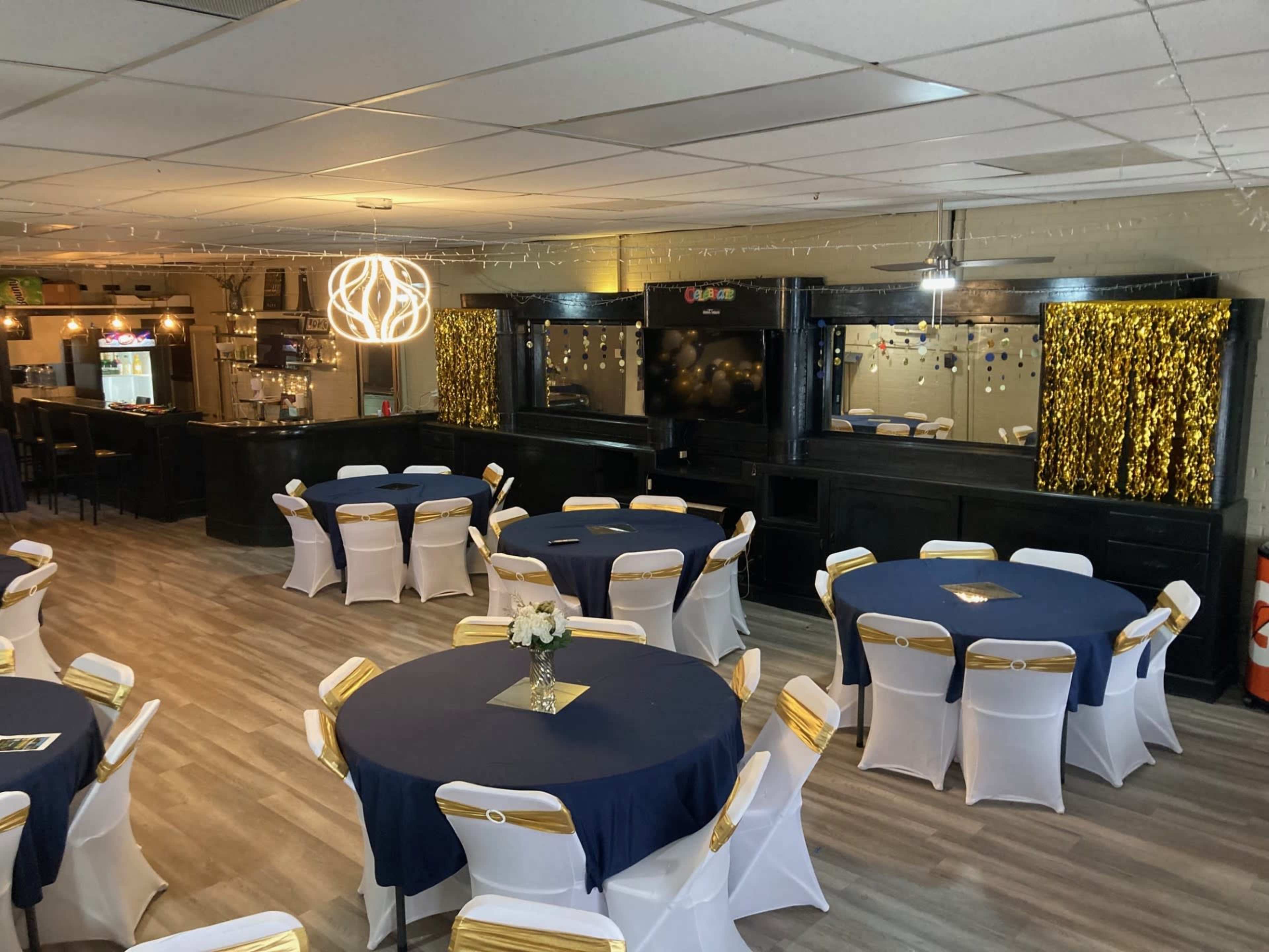 A spacious event hall with round tables set with navy blue tablecloths and white chairs adorned with gold sashes, featuring a bar area in the background and decorative lighting.