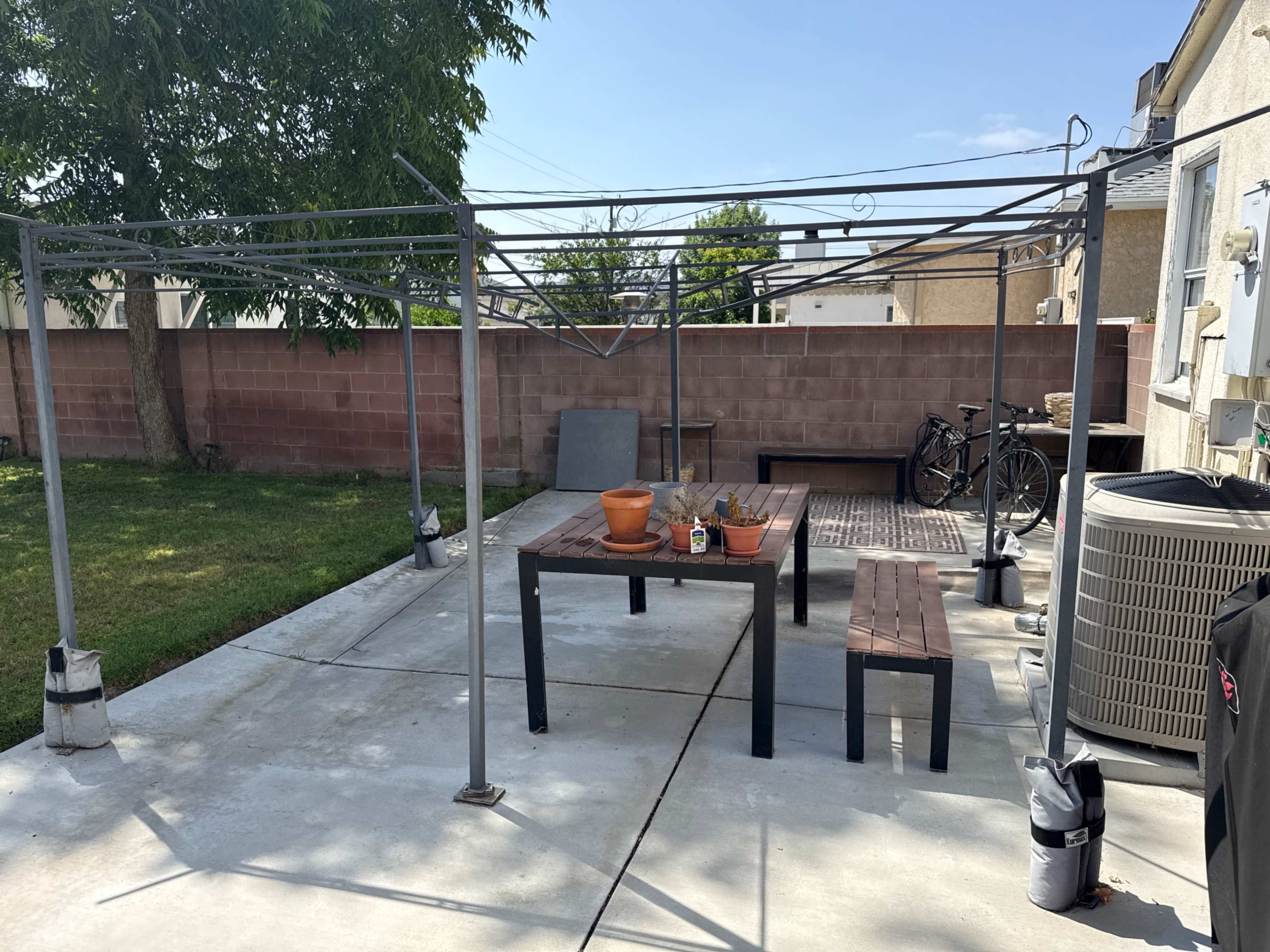 The image shows a backyard with a metal frame structure, a wooden table and benches, a potted plant, and a bicycle leaning against a wall.