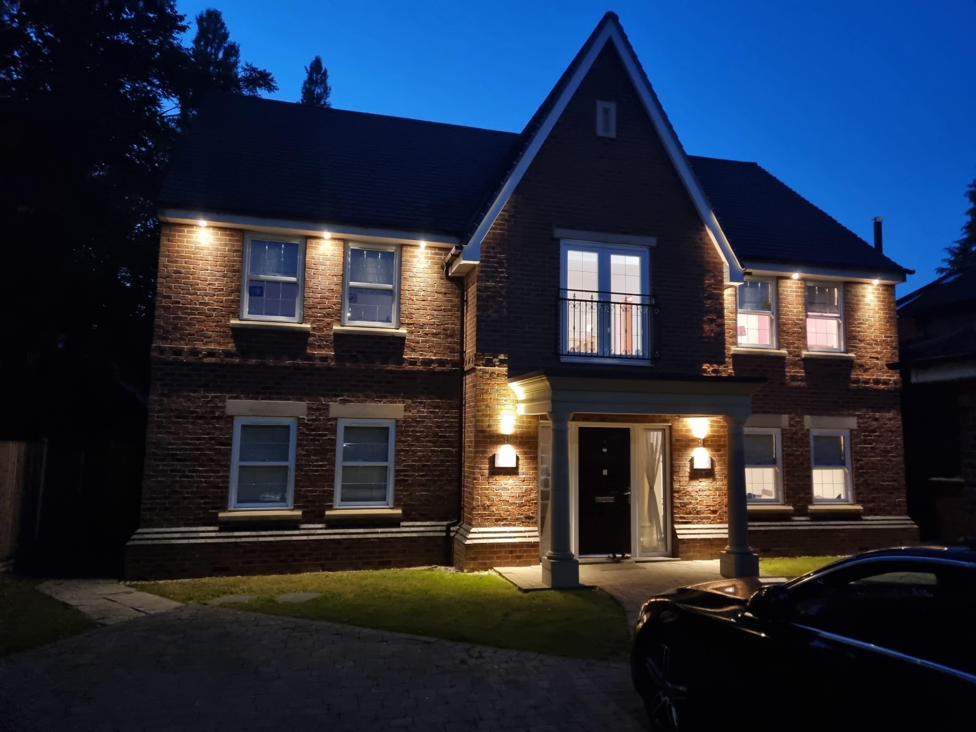 A two-story brick house is illuminated by lights at dusk, featuring multiple windows and a central front door.