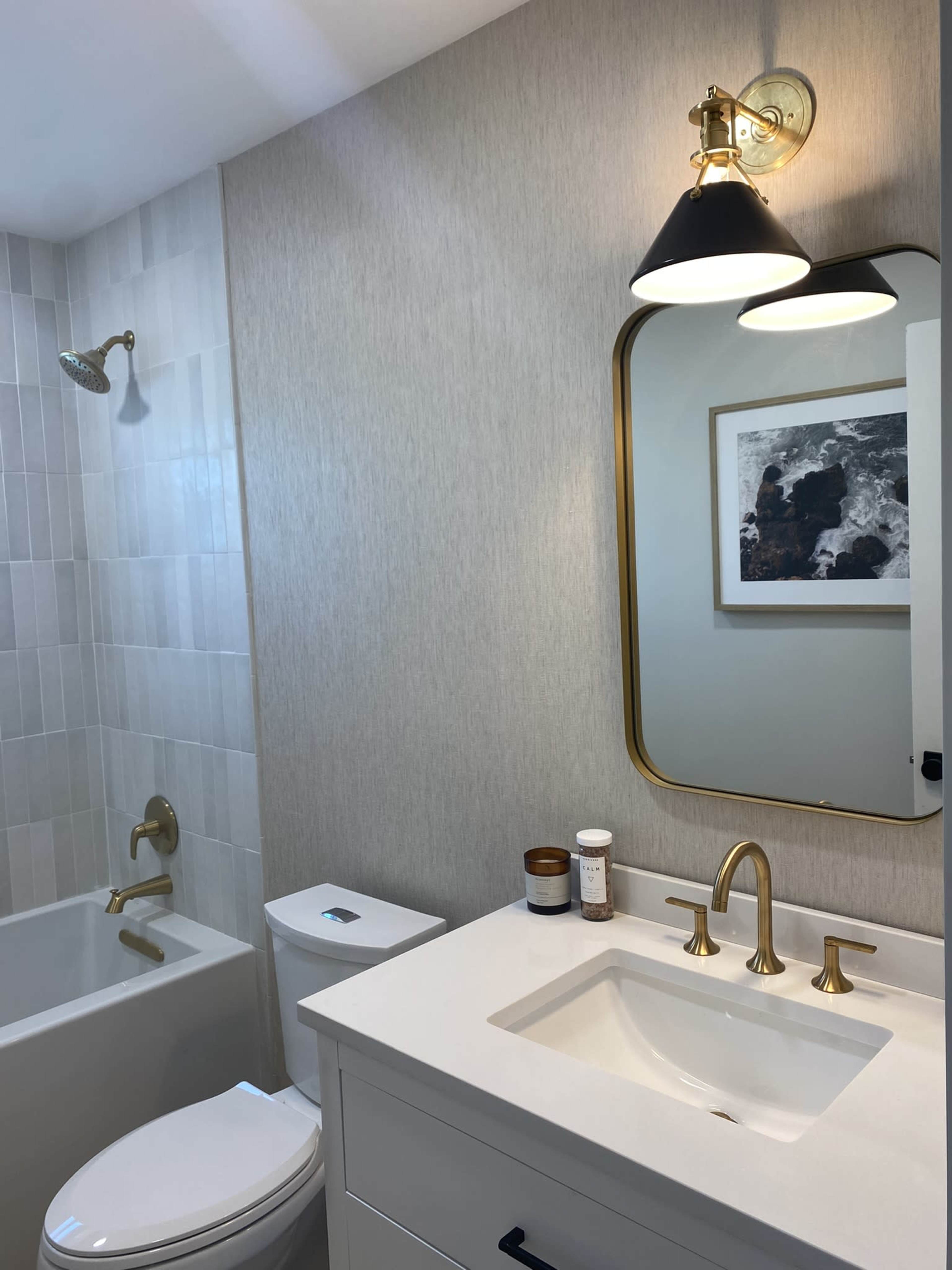 The image shows a modern bathroom featuring a white vanity with gold fixtures, a large mirror, a bathtub, and a showerhead, all against a light-colored wall.