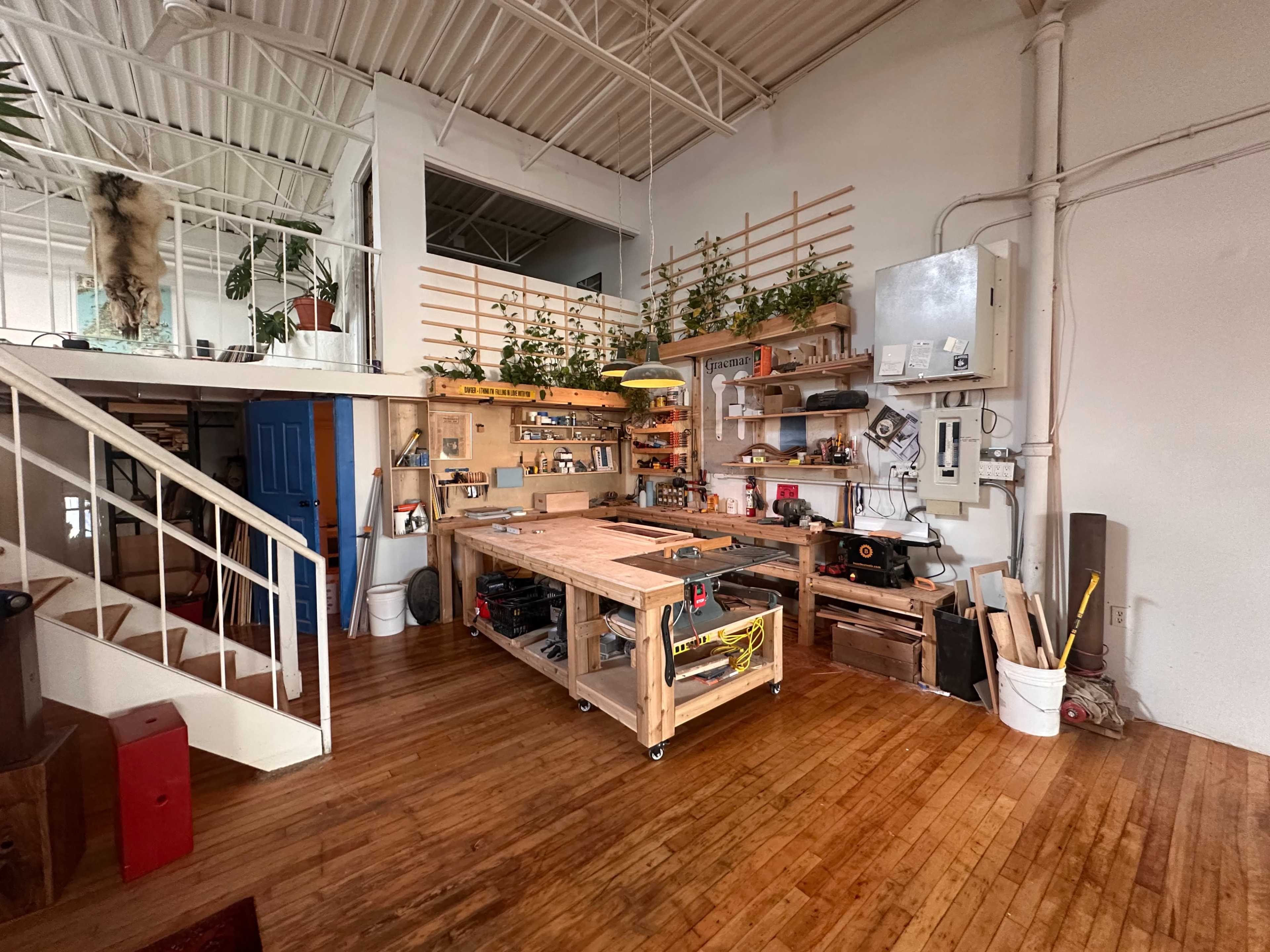 The image shows a woodworking workshop with a central workbench, tools neatly arranged on shelves, and plants placed along the walls.