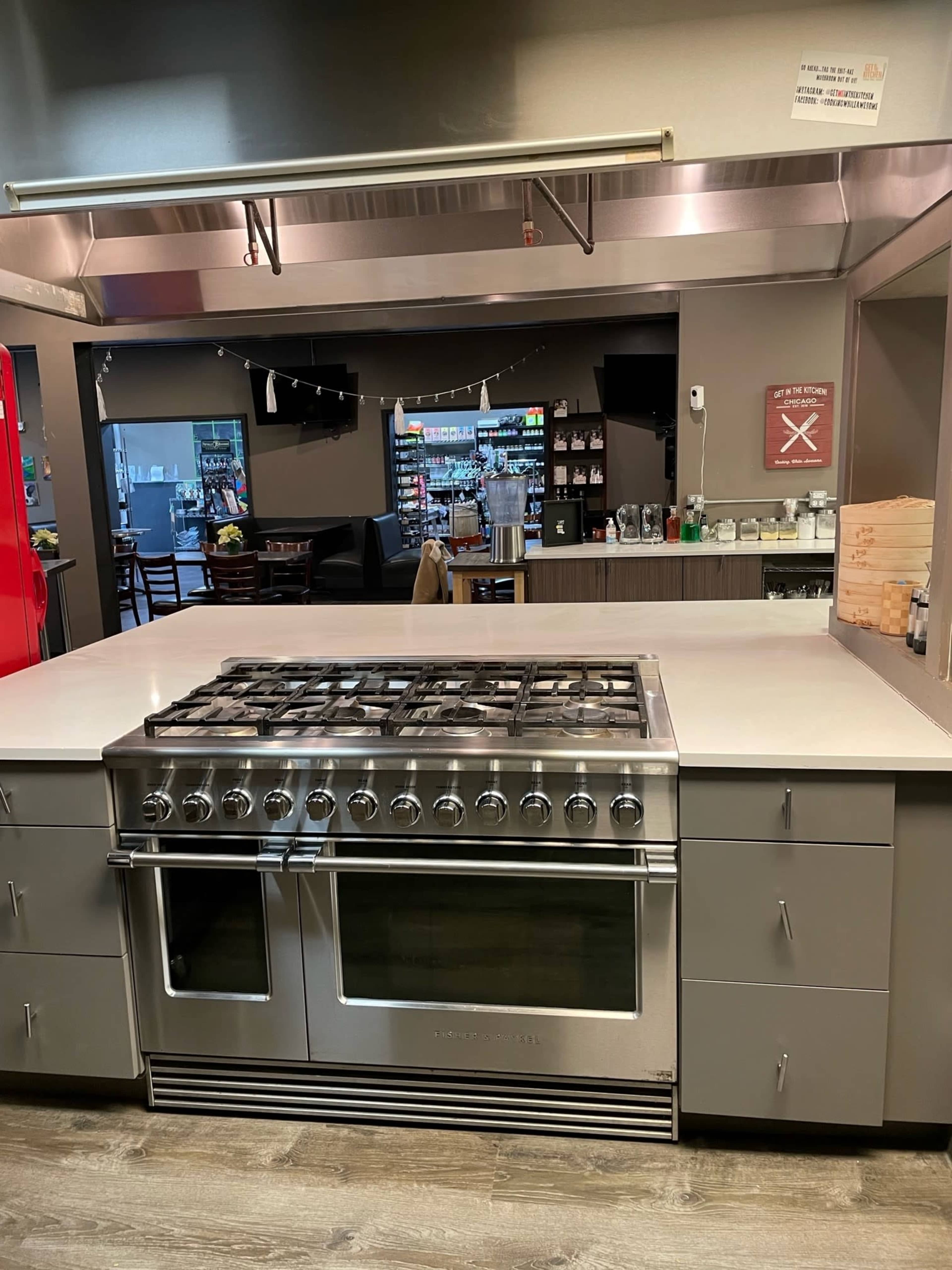 The image shows a modern kitchen with a large stainless steel gas range on an island countertop and a background featuring a dining area and shelves of supplies.