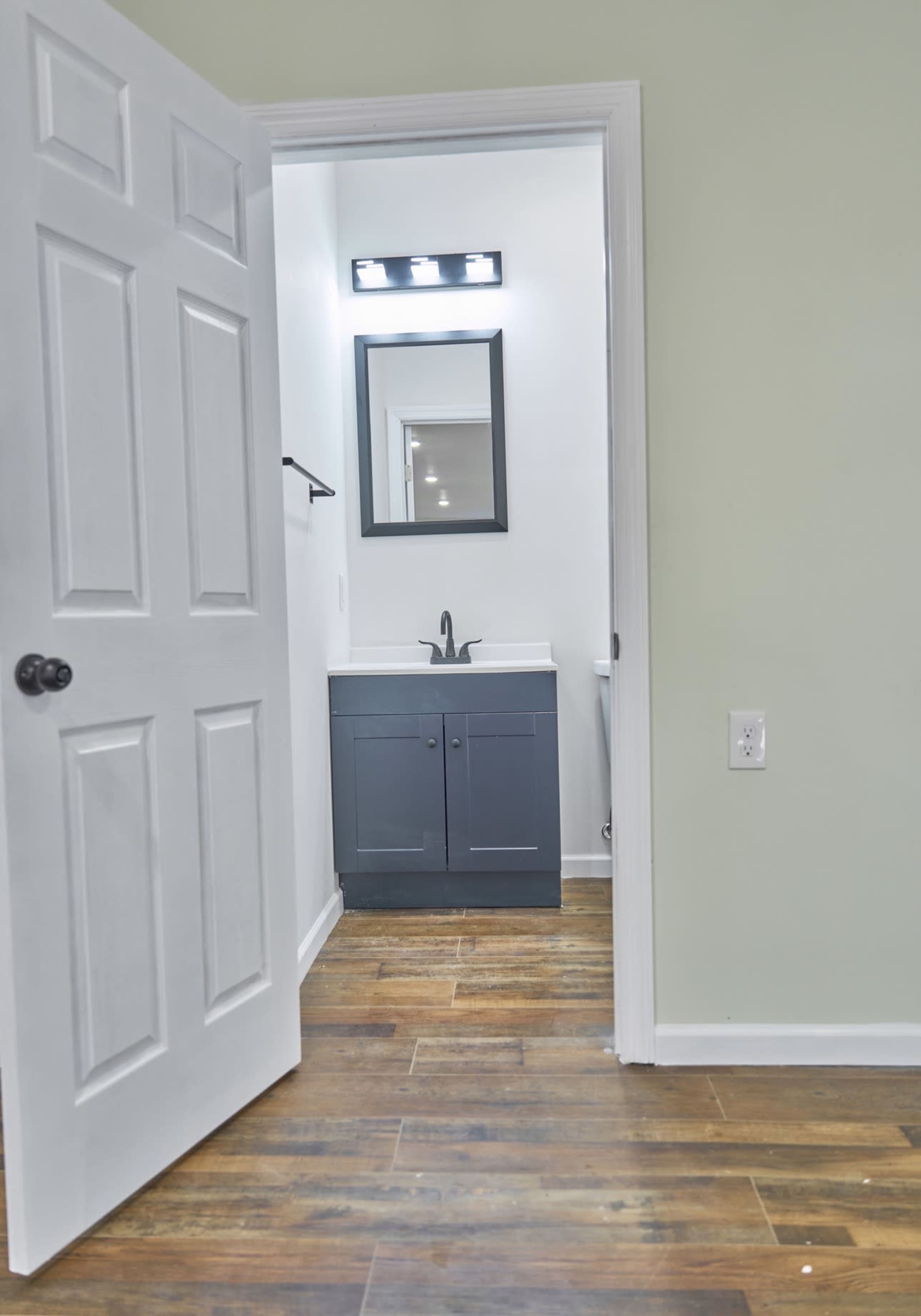 The image shows a bathroom with a gray vanity, a large mirror above it, and a door partially open, revealing wood-like flooring.