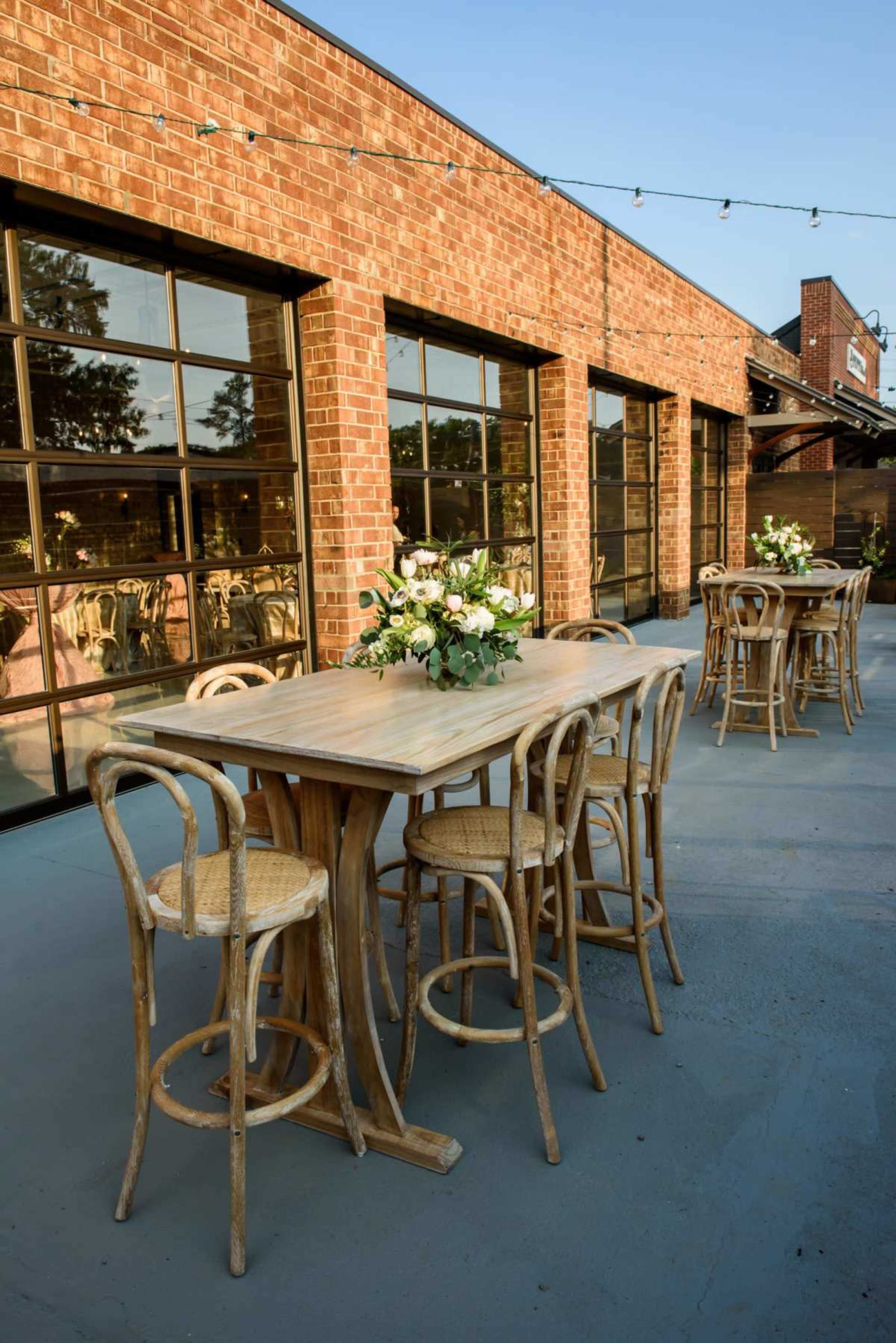 A wooden outdoor dining area features a long table with chairs and floral centerpieces against a backdrop of a brick building and large windows.