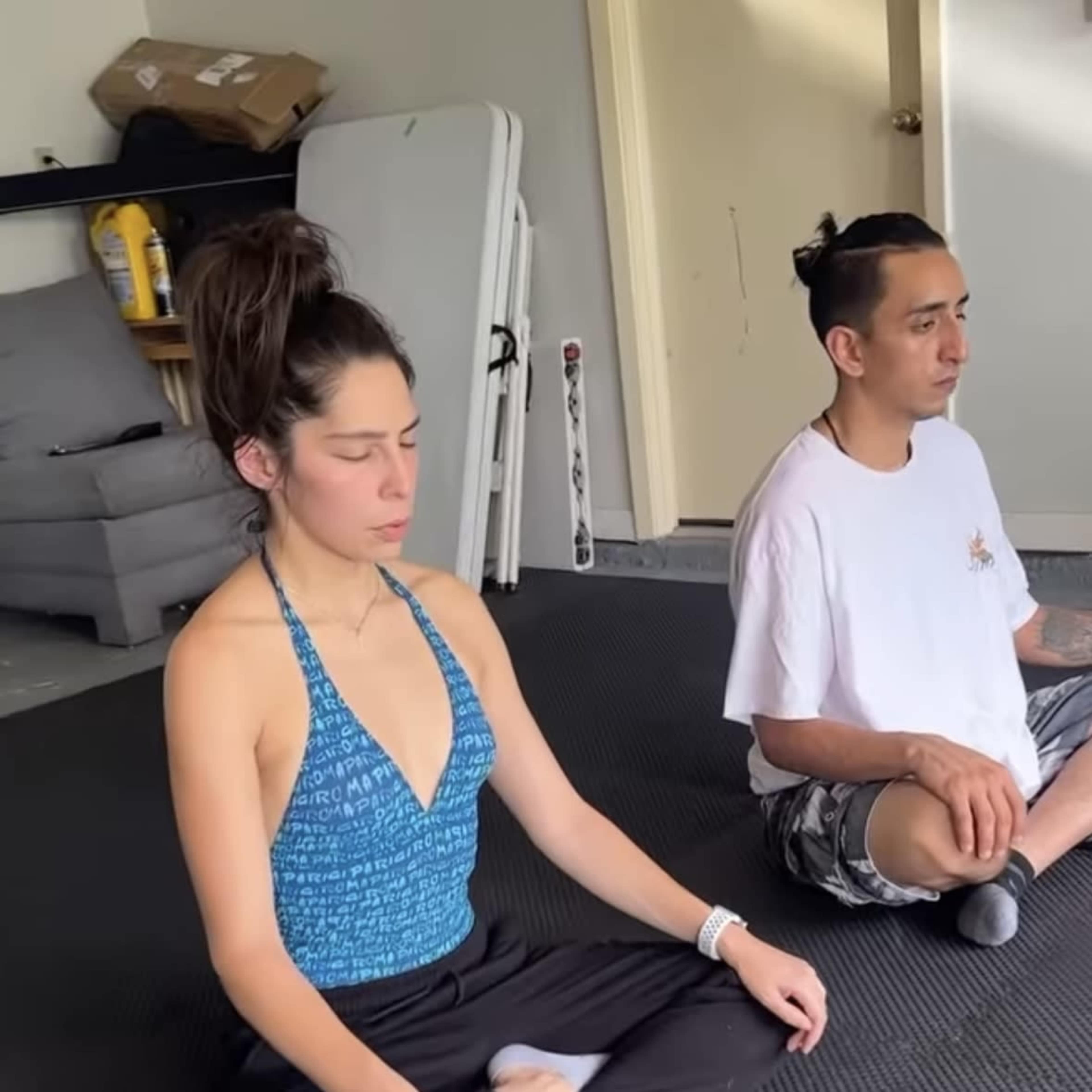 Two individuals sit cross-legged on a black mat in a calm indoor space, with their eyes closed in meditation.