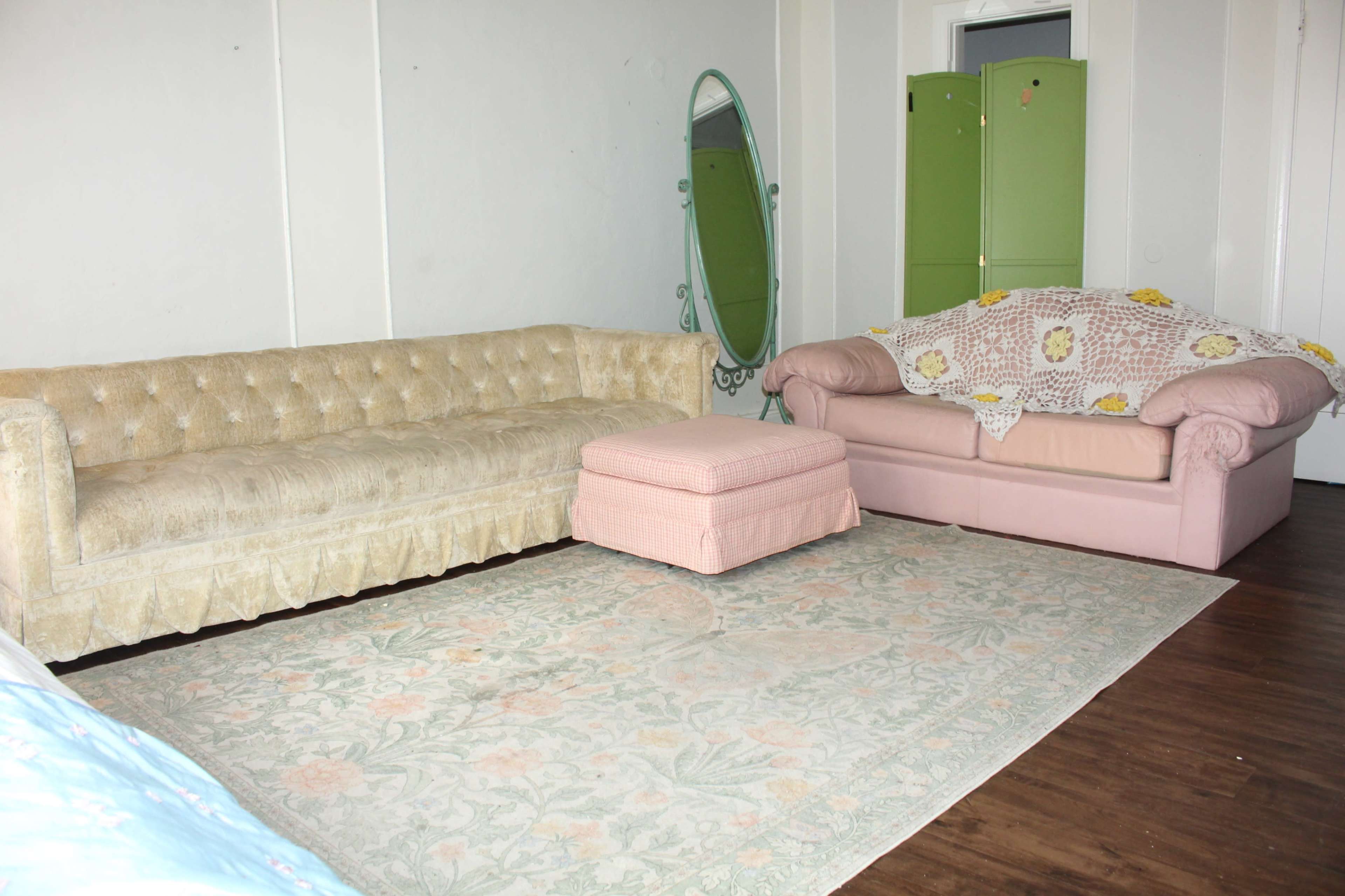 The scene depicts a living room with a light beige tufted sofa, a pink sofa, a pink ottoman, and a floral area rug, alongside an ornate green mirror and a green folding screen.