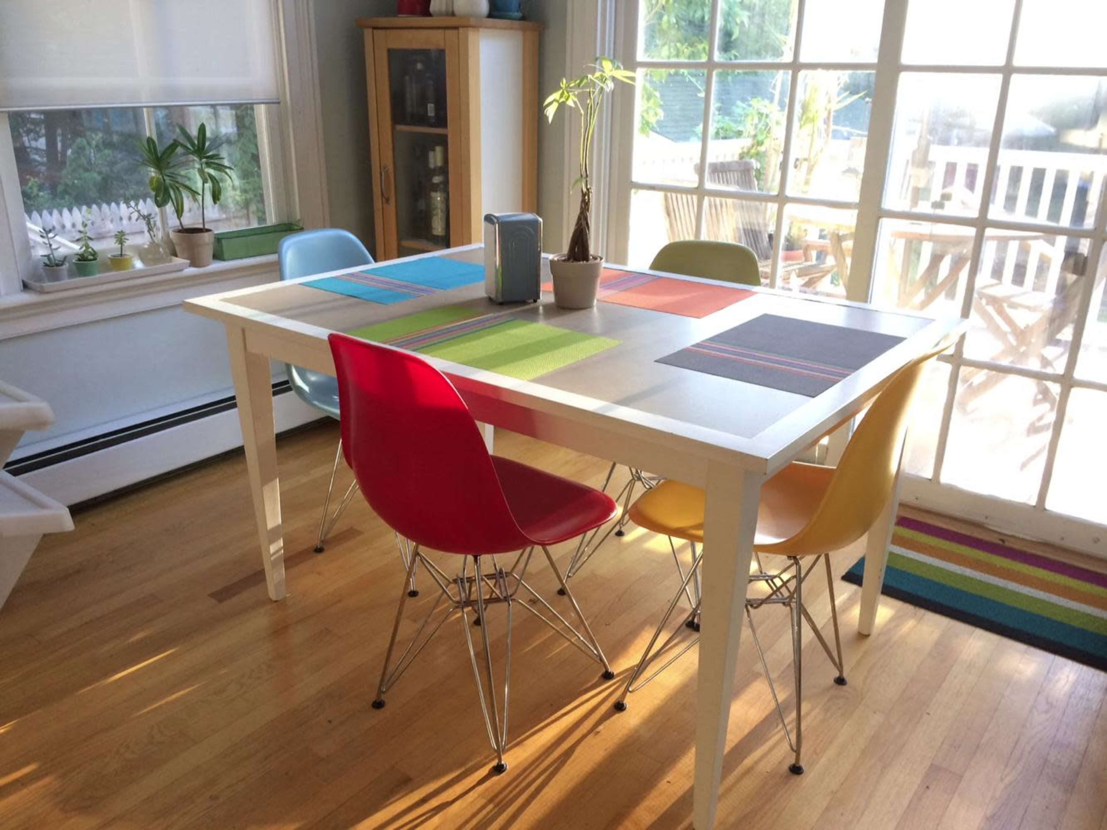 A dining table with four colorful chairs is set in a well-lit room, surrounded by plants and a large window.