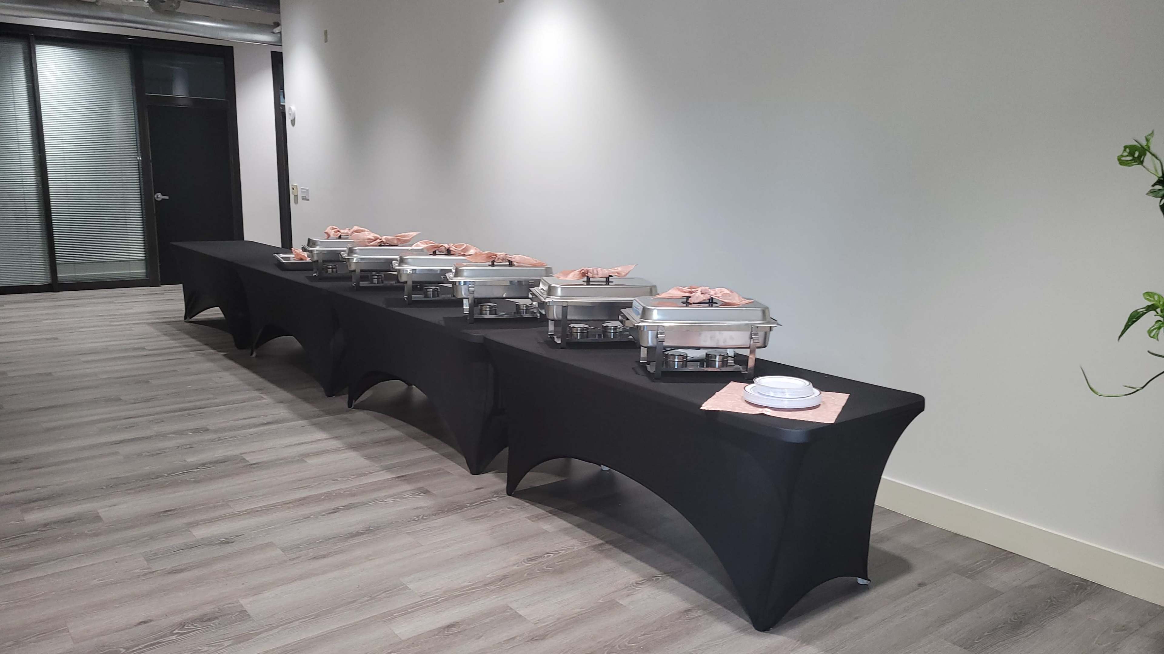 A row of chafing dishes with food covers is set up on black tablecloths in a modern hallway.