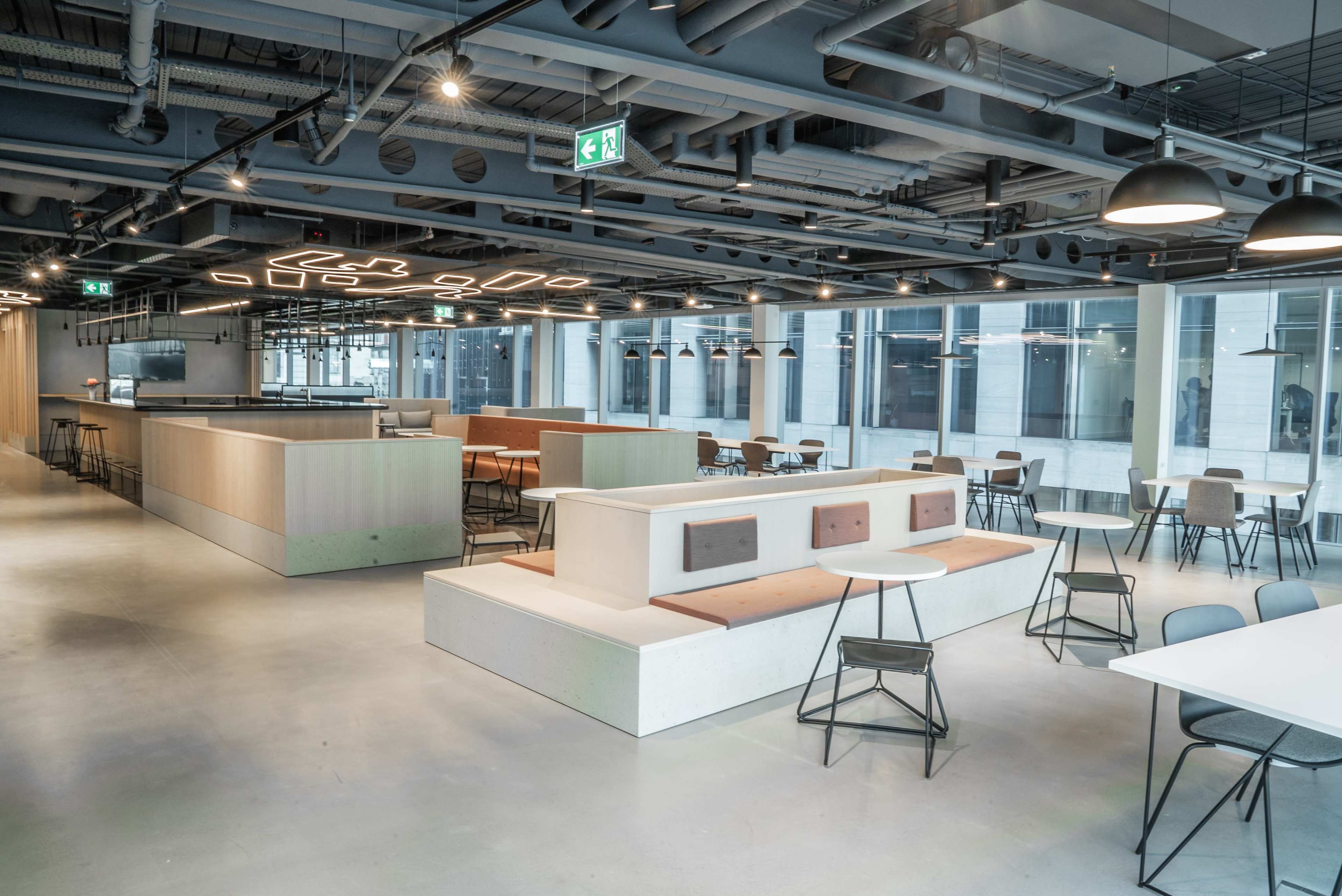 The image shows a modern office space with an open layout featuring a café area, seating arrangements, and large windows providing natural light.