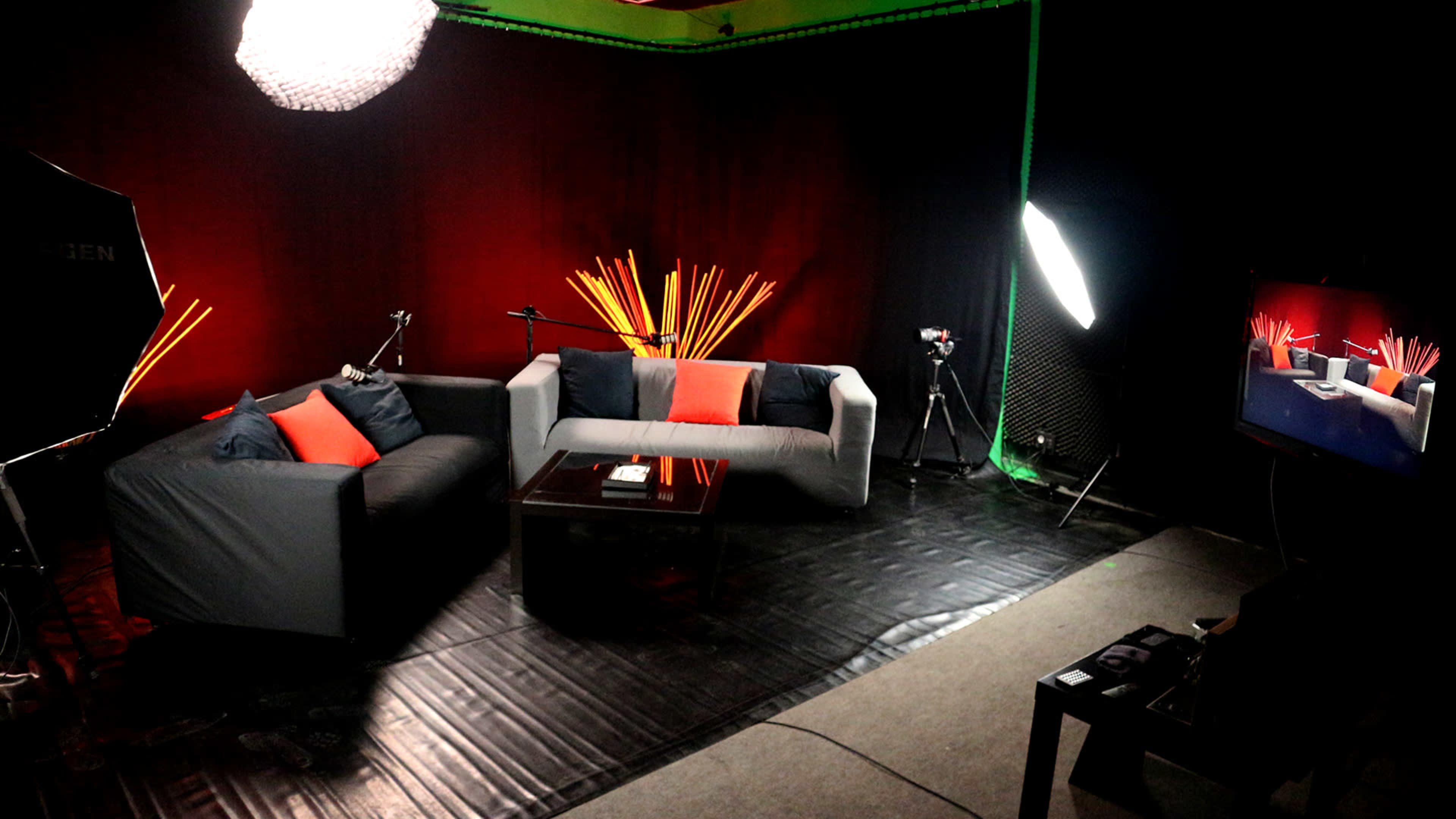 The image shows a modern studio setup with two gray couches, decorative orange pillows, a coffee table, and lighting equipment arranged against a dark backdrop.