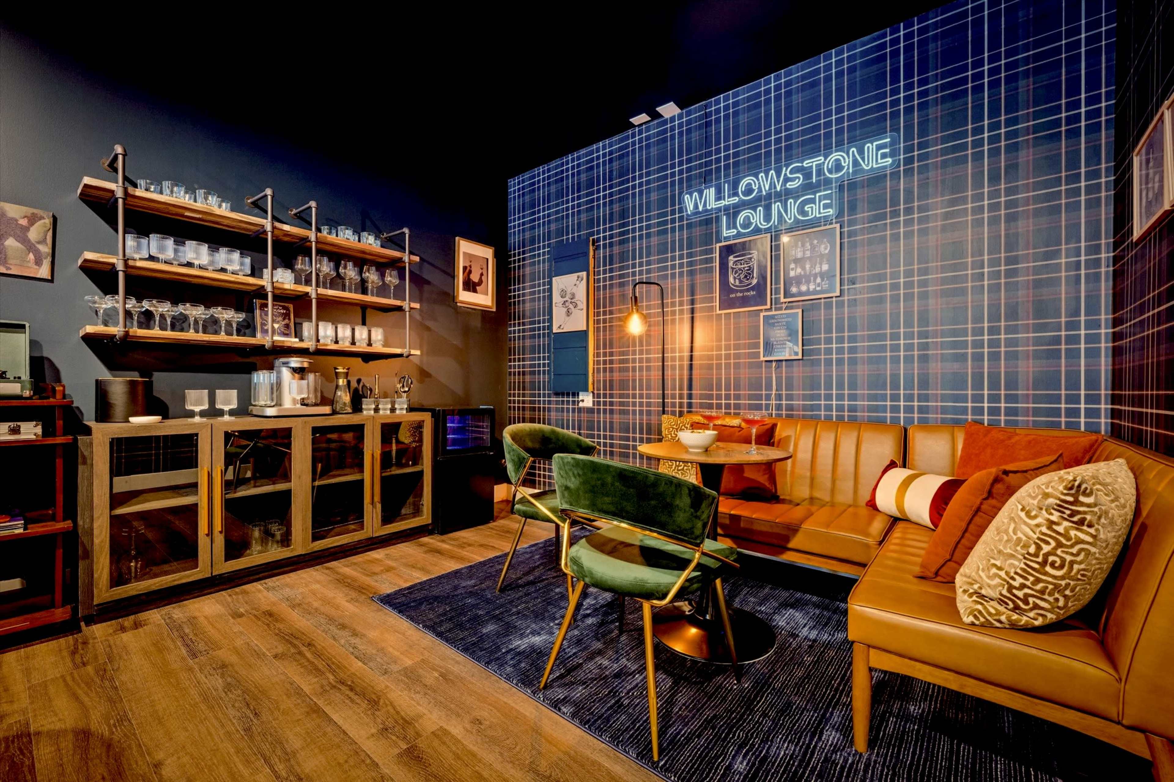 The image depicts a cozy lounge area featuring a brown leather sofa, green chairs, a round table, and a bar unit with glassware against a plaid accent wall.