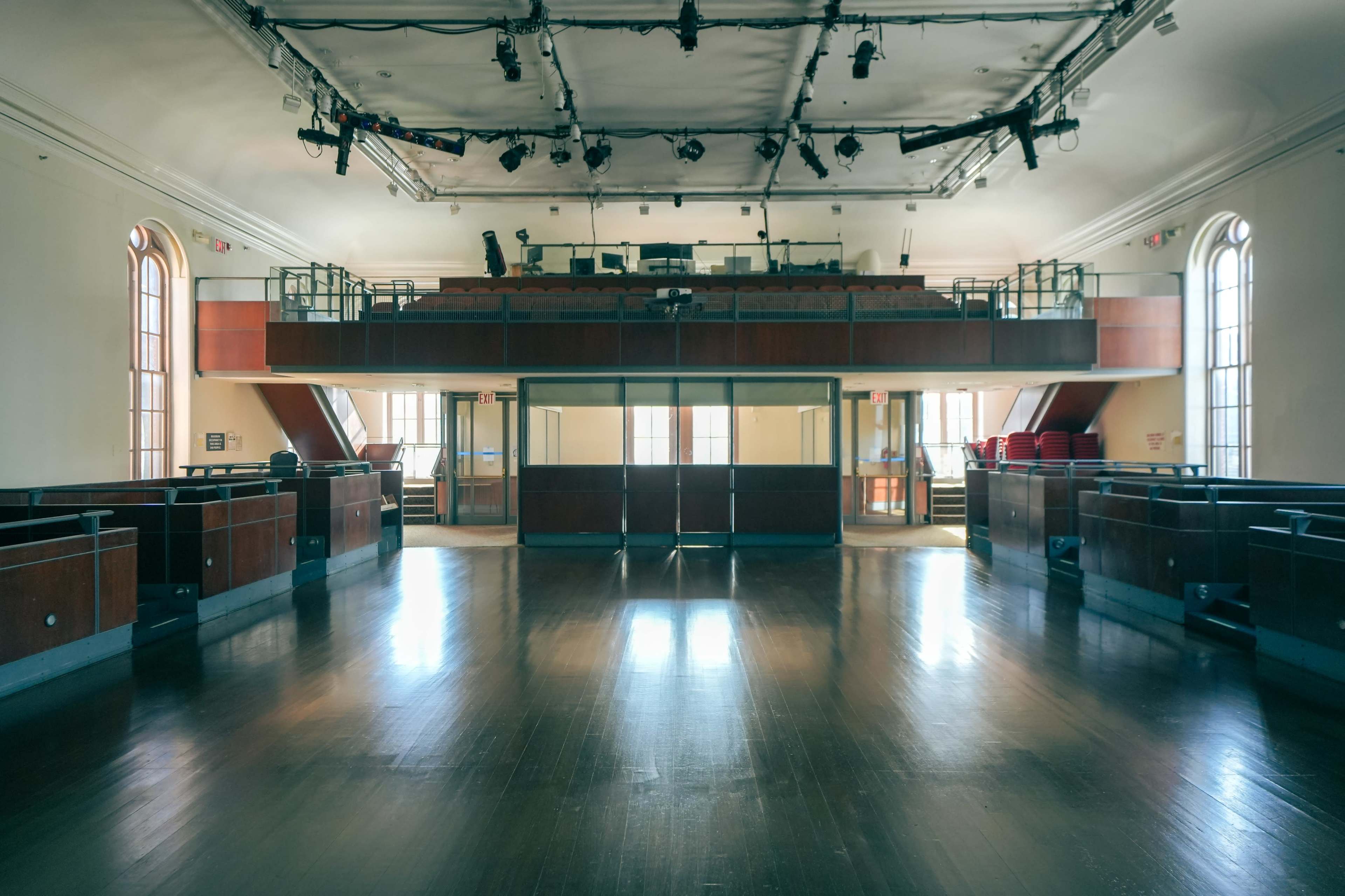 An empty theater with a raised balcony, wooden flooring, and large windows.